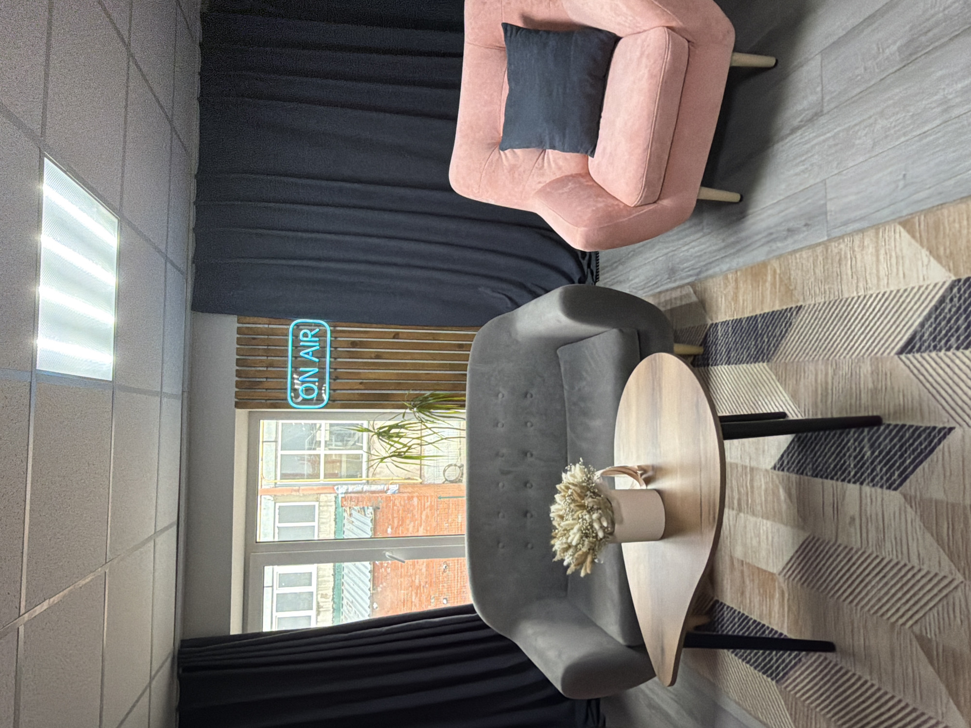 The image depicts an interior scene, possibly a studio or office, in what is presumed to be Chaikovsky, Russia based on location information. A window reveals a view of what appears to be an urban residential area, with brick buildings and multiple windows. A bright neon sign reading "ON AIR" is visible.
The room has a contemporary design with a pink armchair featuring a dark-colored pillow, a gray modern-style sofa, and a uniquely shaped wooden coffee table with a vase holding dried floral arrangements. A patterned rug is visible on the floor. Dark curtains provide privacy. The walls are white and appear to be made of square panels. The room seems set up for a podcast or recording session, given the "ON AIR" sign. No people are present in the photograph.