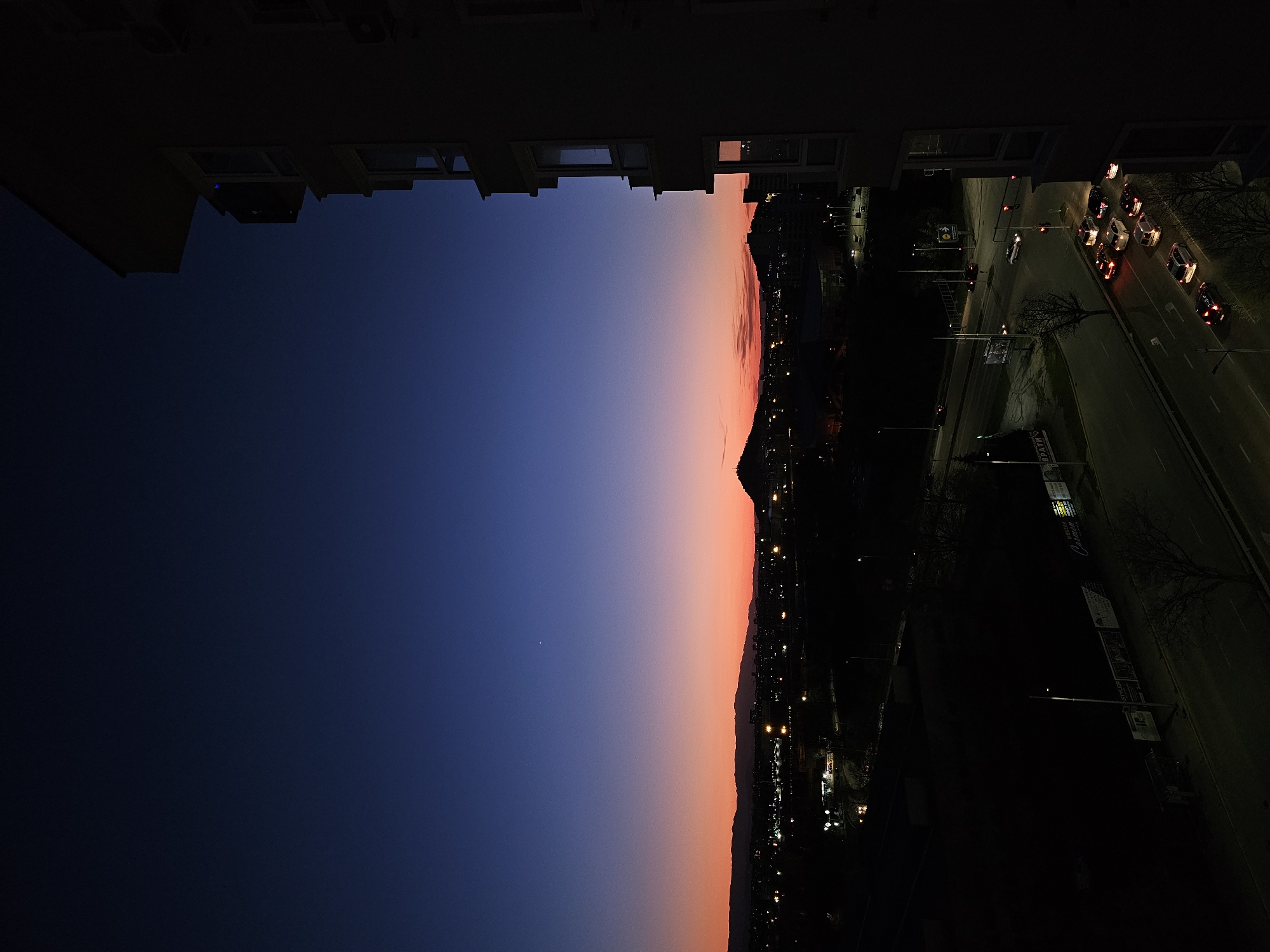 The image shows a view of Plovdiv, Bulgaria at dusk. The perspective is from a high vantage point, likely an upper floor of a building, looking towards a horizon with a gradient sky transitioning from dark blue to orange. The silhouette of the Rhodope Mountains is visible along the horizon line. Below the mountains, city lights illuminate a valley and roads, including traffic with car headlights and tail lights. Sections of a building frame the top and bottom of the view. The buildings appear to be residential apartment blocks.