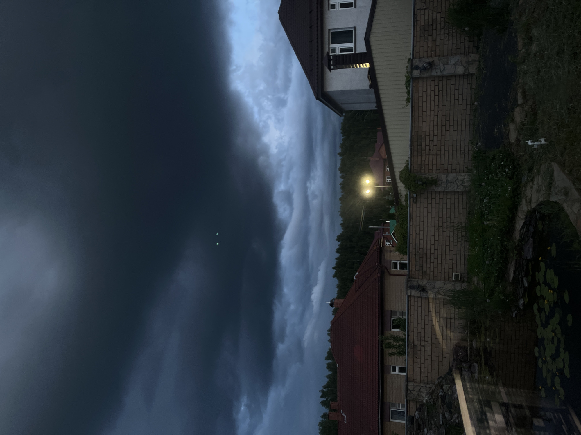 The image depicts a rural or suburban residential scene at dusk or night, set against a dramatic sky dominated by heavy, dark storm clouds on the left and more textured, lighter clouds to the right. The overall impression suggests an impending or recent storm.
On the right side of the frame, two houses are visible. The upper house features light-colored siding or stucco, dark window frames, and a dark, steeply pitched roof. The lower house has brick walls with a red tile roof. Between these structures, a bright, warm-toned outdoor lantern or light illuminates the immediate area. A dense line of dark forest trees forms the horizon in the middle ground.
In the foreground, near the base of the lighter-colored house, a white drone is resting on the grassy ground. Further to the bottom right, a pond or water feature is visible, covered with numerous lily pads, some bearing small yellow flowers. The ground around the houses and pond is covered in grass and some wild foliage, with visible stone and brick retaining walls. Two small, faint green light points are discernible high in the dark expanse of the sky on the upper left, distinct from the grounded drone.
There are no people visible. The scene conveys a quiet, somewhat tense atmosphere due to the formidable clouds. The location is contextualized as Ореховка, Russia. No visible text is present in the image.