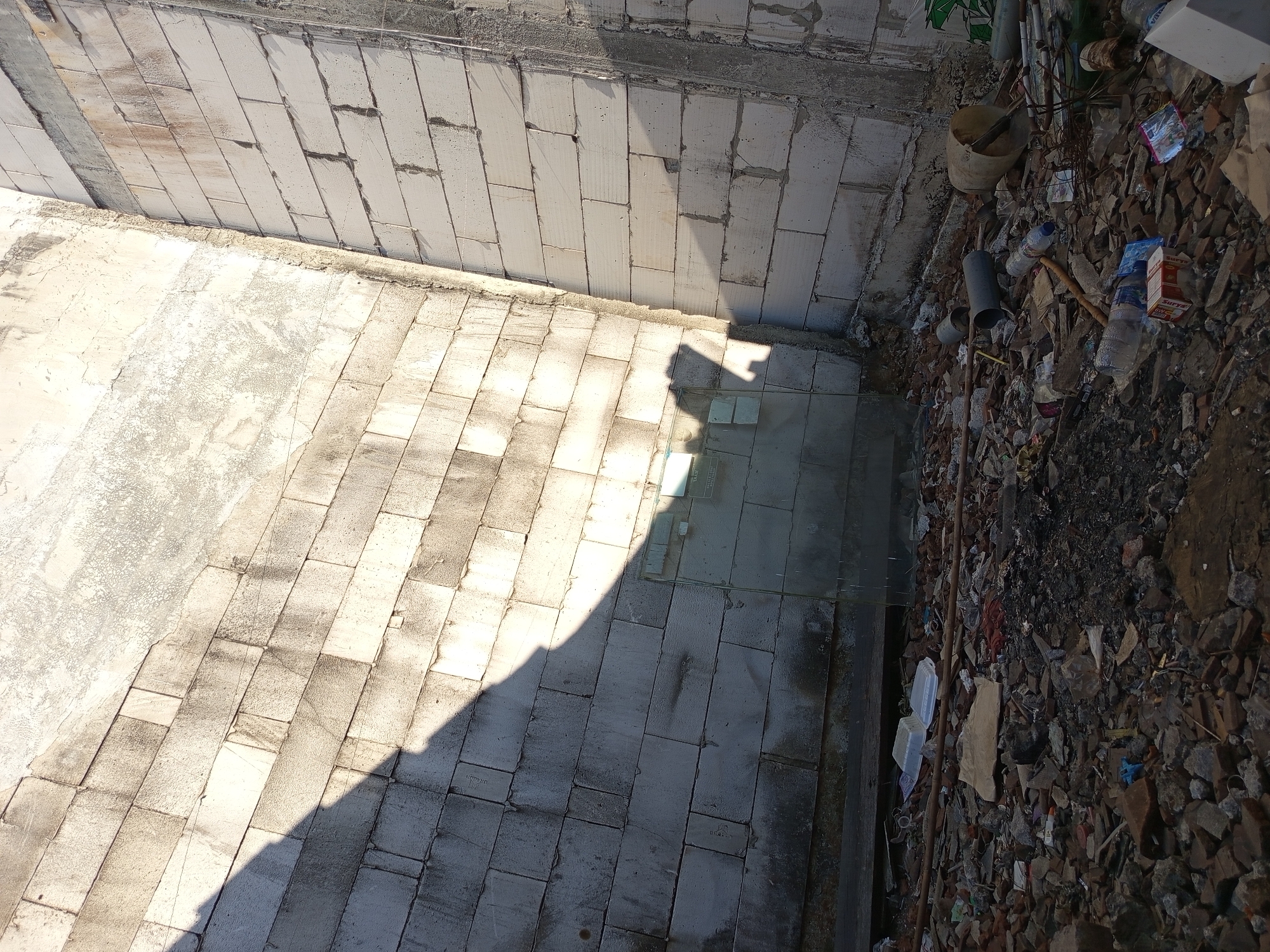 The image depicts a construction site in Tebel, Indonesia, littered with debris. The foreground is a chaotic mix of rubble, broken bricks, plastic bottles, cardboard packaging, and other discarded materials. In the background, two unfinished brick walls form a corner, casting a long shadow from an unseen object. The walls are constructed of light-colored cinder blocks, some showing signs of wear and discoloration. The overall impression is one of neglect and disarray.