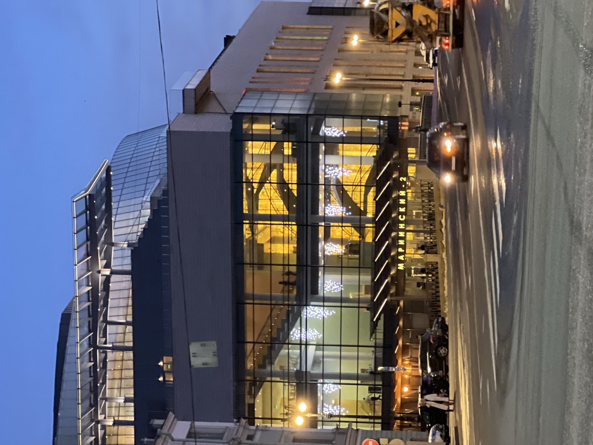 The image shows a modern building, with the words "Мариинский-2" (Mariinsky-2) visible on its facade, indicating the location is likely in Saint Petersburg, Russia, specifically the Mariinsky Theatre's second stage building. The architecture features a combination of glass and solid surfaces, with multiple levels and internal staircases visible through the glass facade. Christmas decorations, in the form of lit garlands or similar arrangements, are visible within the building. The exterior shows signs of nighttime or dusk, with artificial lighting illuminating the building. Streetlights are present along the road, and there's evidence of a wet road surface reflecting light. A few people are standing on the sidewalk in front of the building and cars are parked along the curb. A car with headlights is driving on the street. A construction crane or similar machinery is visible in the background.