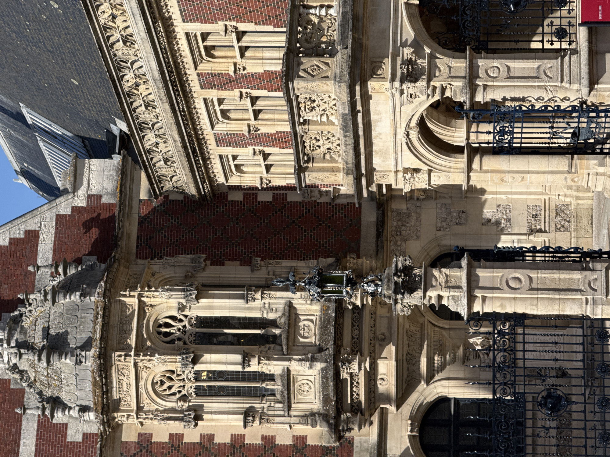 The image captures a detailed view of an ornate, historic building from a low, upward-looking perspective, under bright, clear daylight. The architecture combines striking red brickwork, often laid in patterns, with extensive sections of light-colored, intricately carved stone.
The building exhibits characteristics of Gothic or Neo-Gothic design, featuring elaborate stone tracery around windows, pointed arches, and decorative cornices. A steep, dark slate roof with dormer windows is visible against a clear blue sky in the upper left. The facade is adorned with a variety of sculpted elements, including floral and foliate motifs, geometric patterns, and grotesque figures. One prominent detail is a large, winged creature, possibly a dragon or griffin, extending from the facade, appearing to hold a lantern or escutcheon.
Several windows with dark or stained glass are visible, some rectangular with circular elements, others with more complex curvilinear tracery. Wrought-iron railings with decorative scrollwork and finials protect what appear to be balconies or openings on various levels. Strong shadows cast across the facade emphasize the intricate three-dimensional relief of the carvings and architectural features, indicating a sunny day.
No people are visible, and no specific activity is depicted. A red sign with white text, partially visible behind a wrought-iron gate, clearly displays the word "DISTILLERIE". This sign serves as a significant location cue, strongly suggesting the building's historical or current function. The building's grandeur and detailed craftsmanship dominate the scene.