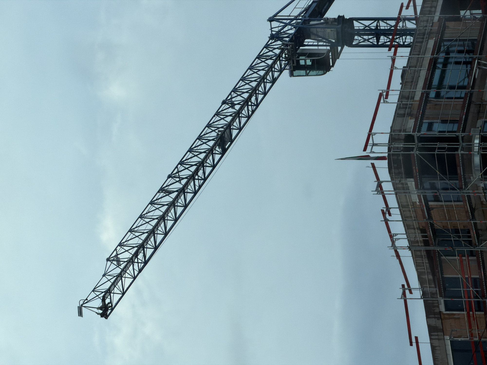 A crane is shown adjacent to a building under construction in Sofia, Bulgaria. The crane is a dark gray lattice structure with a cab at the top and a jib extending to the left. The building is in the process of being built, with scaffolding visible along its side. The background is a cloudy sky.