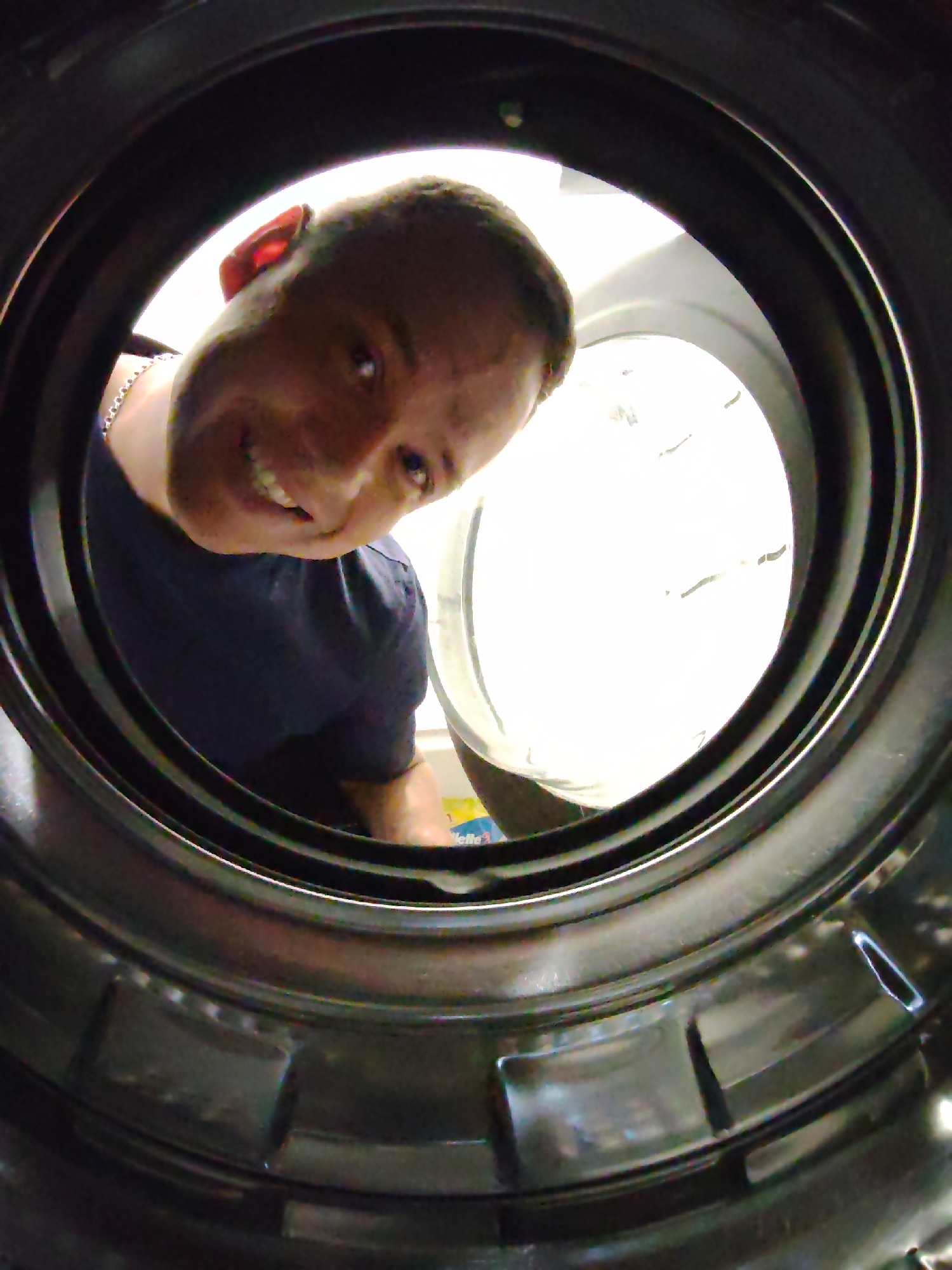 A man looks into a washing machine from the interior of the drum. The man is fair-skinned with short hair, and is wearing a dark blue t-shirt and a silver chain. The drum is a metallic grey with a pattern of rectangular insets. The inside of the machine is brightly lit. The image was taken with a wide-angle lens and is presented from the perspective of a viewer inside the drum. The location is unknown and there is no evidence of Chaikovsky, Russia.