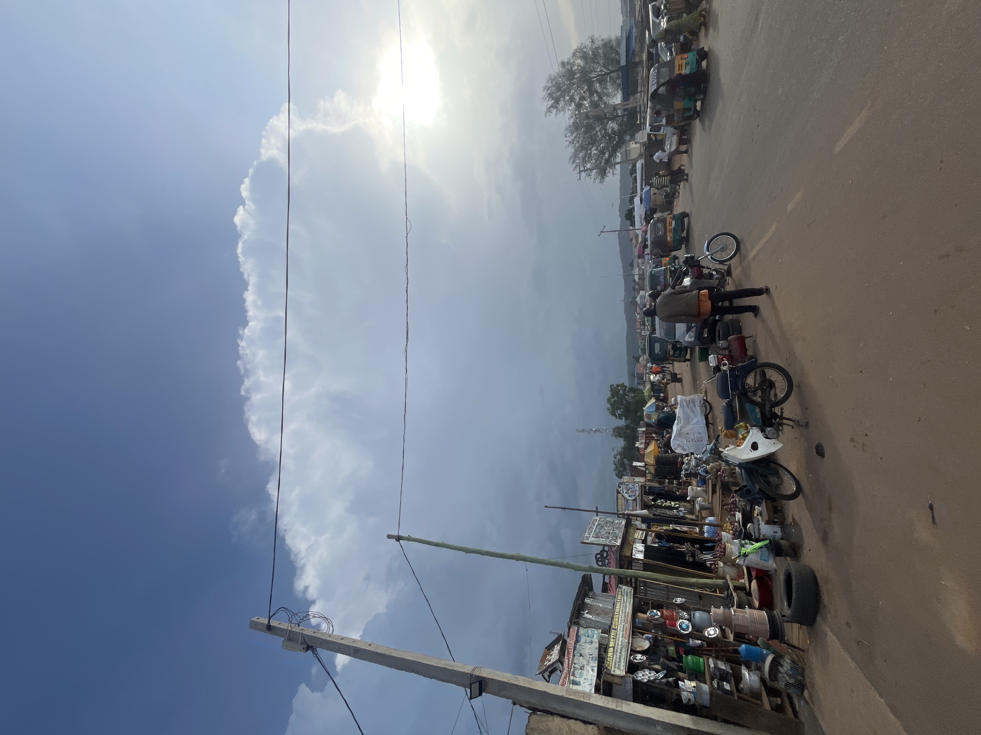 This image depicts a bustling street market scene in Rafin Par, Nigeria. The foreground shows a dirt road with a few parked motorcycles and scattered debris. To the left, market stalls are set up along the roadside, displaying various goods such as metal products, containers, and what appear to be food items. People are seen milling about the market and on the road, some walking and others interacting with vendors. In the background, a line of vehicles, including what look like tuk-tuks and cars, are moving along the paved road, indicating ongoing commerce and transportation. The sky is partly cloudy with a bright sun peeking through, suggesting it is daytime. Power lines and poles are visible, crisscrossing the scene and indicating the presence of electrical infrastructure. The overall impression is one of a lively, active marketplace in an urban or semi-urban setting.