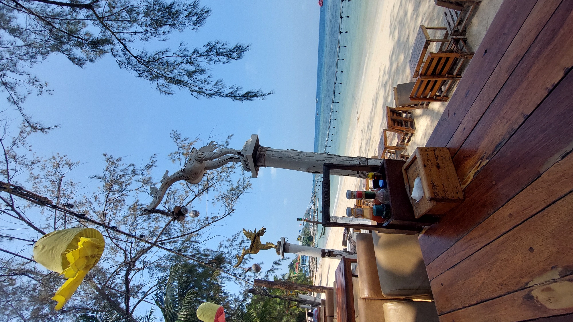 The image depicts a beach scene in Koh Tao, Thailand. A wooden table occupies the foreground, with condiments and a tissue box placed on it. Beyond the table, wooden chairs are arranged on the sandy beach. In the midground, the beach meets the turquoise sea, with a string of lights stretching along the shoreline. Decorative poles with bird sculptures are visible among the trees. The sky is clear and blue, with tree branches extending into the upper portion of the frame. A yellow ribbon hangs from the branches.