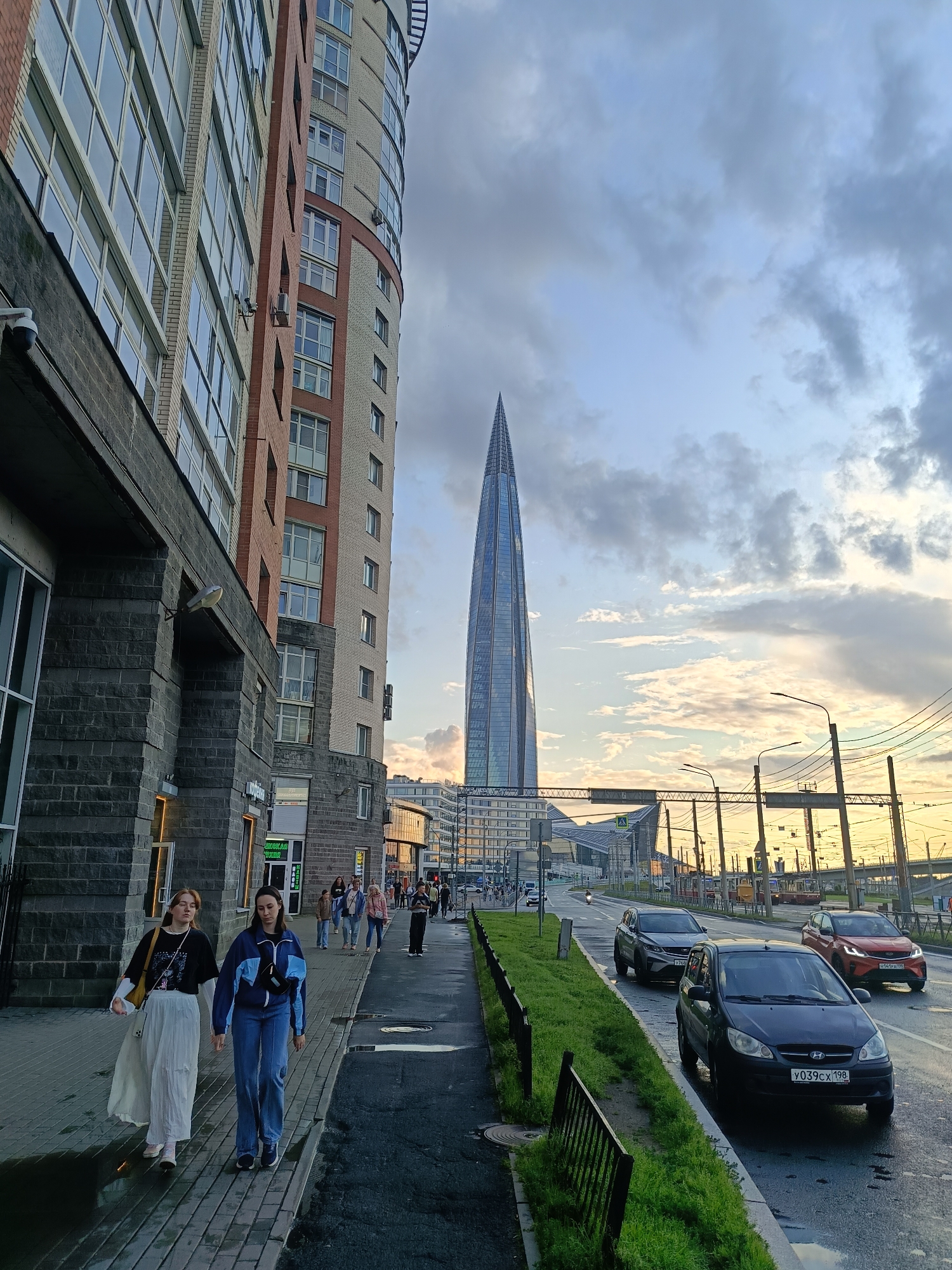 The image depicts a street scene in Saint Petersburg, Russia, likely on the Vasilyevsky Island, with the Lakhta Center skyscraper visible in the distance. The foreground features a modern brick and stone building with large windows, and a sidewalk where two young women are walking. They are wearing casual clothing, with one in a black t-shirt and white skirt, and the other in a blue tracksuit and jeans. Other pedestrians are seen further down the sidewalk, and cars are driving on the wet asphalt road to the right. The sky is cloudy with hints of sunset colors. The overall atmosphere suggests an urban environment with contemporary architecture.