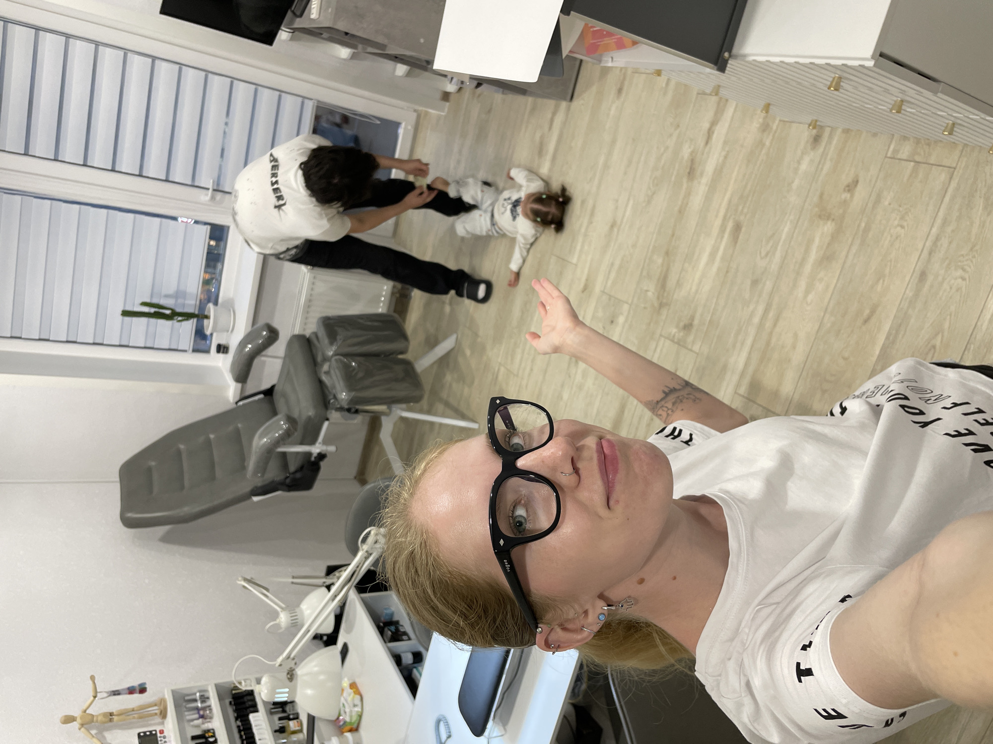 The image shows an interior scene, possibly a home, in Izhevsk, Russia. The foreground is dominated by a woman taking a selfie, wearing glasses and a white T-shirt. In the background, a man in a white shirt and black pants is interacting with a toddler lying on the wooden floor. A gray dental chair and other salon equipment are visible, suggesting the room may be used for professional services. Through the window, other buildings are seen, indicating the location is likely within a residential area of Izhevsk.