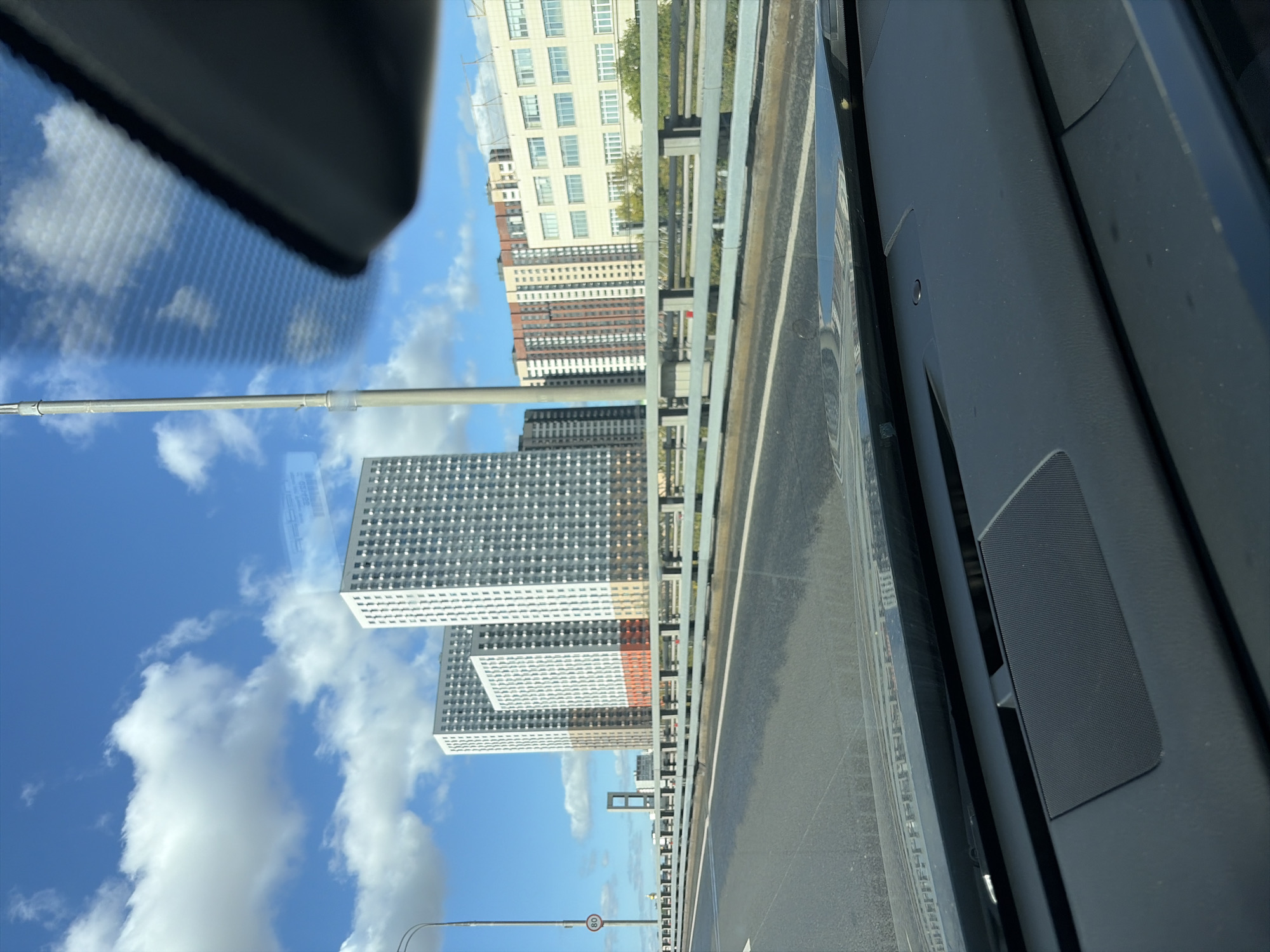 The image shows a view from inside a car, looking out at a cityscape. The perspective is from the driver's seat, with the dashboard visible in the foreground. The car is on a highway or elevated road, with a grey metal barrier separating the road from a green area. In the background, several modern apartment buildings are visible. The tallest buildings are white with many windows, and some have colored sections at the bottom. Other buildings in the background are a mix of beige, red, and white with multiple floors and windows. The sky is blue with scattered white clouds. A tall, thin light pole is also visible on the left side of the image, with a speed limit sign indicating "80". Based on the architectural style of the buildings, particularly the tall, white residential complexes, and the presence of a light pole that resembles those found in Moscow, the scene is likely located in Moscow, Russia. Specific identifiable landmarks are not present, but the overall impression is of a rapidly developing urban area.