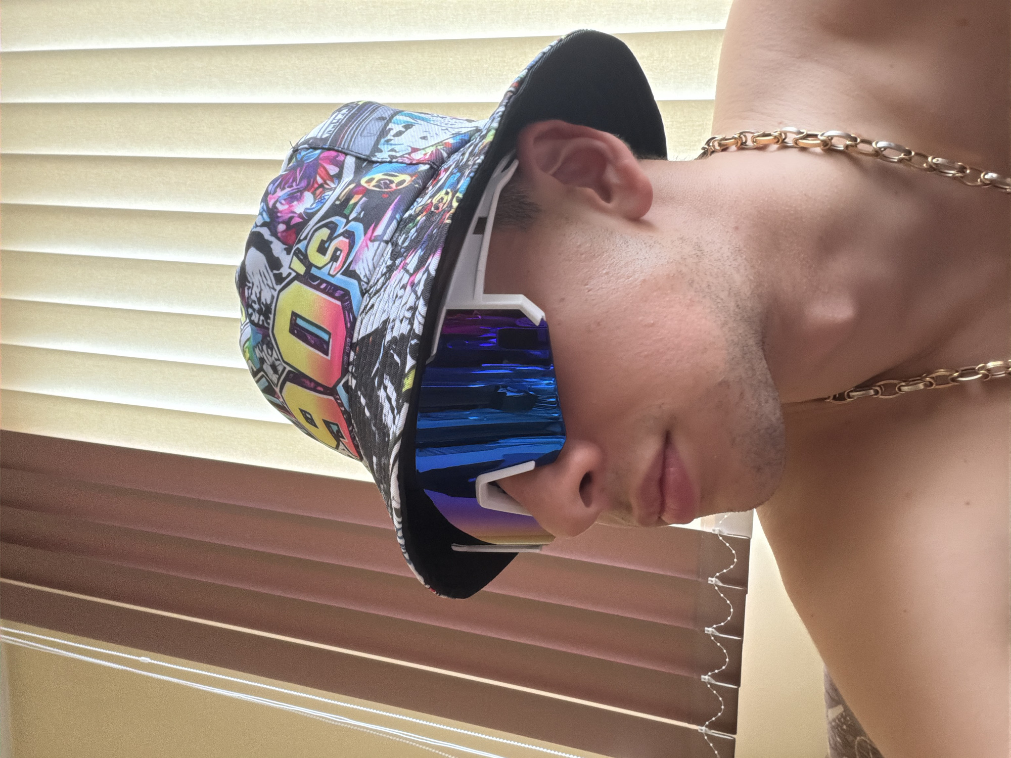 The image shows a person wearing a colorful bucket hat and blue-tinted sunglasses, along with a gold chain around their neck. They are positioned in front of a set of closed Venetian blinds, which are partially visible in the background. The location is unknown, and no specific details about the city are available.