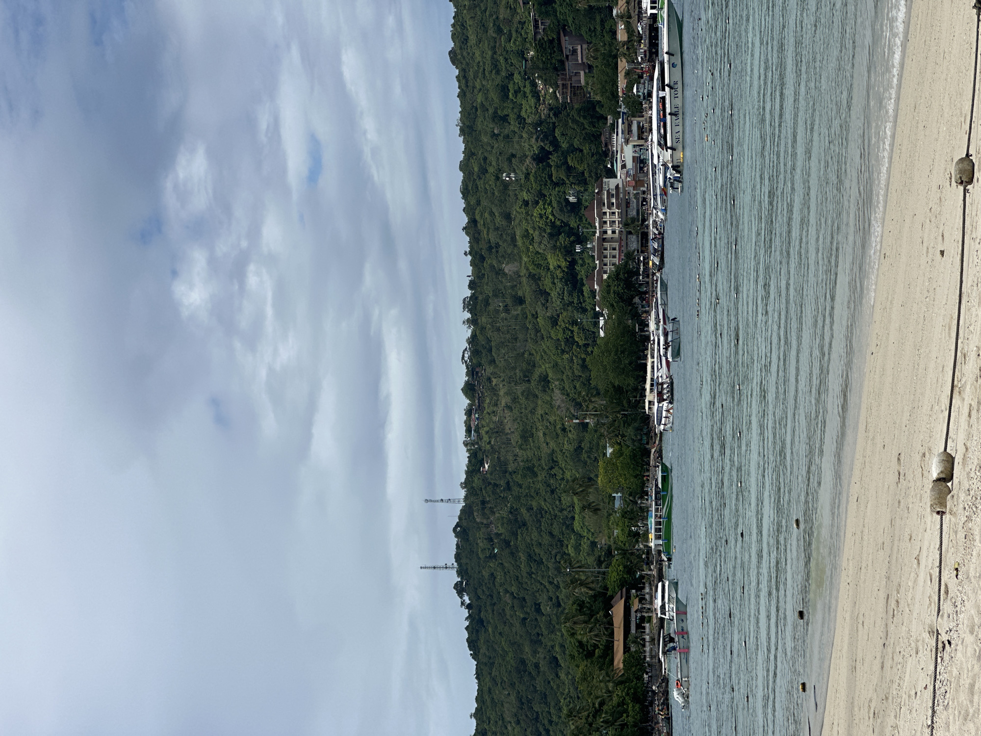 This is a medium-shot, eye-level photograph of a tropical beach scene in Ao Nang, Thailand. The foreground is dominated by a sandy beach with a rough rope secured by buoys. The middle ground features a calm, turquoise body of water filled with numerous boats. These include speedboats, some labeled "SEA EAGLE TOUR," and a larger green tour boat, all anchored or moving slowly. A line of buoys stretches across the water, marking a boundary. In the background, a densely forested, hilly island rises, dotted with buildings and topped by two prominent communication towers. The sky is overcast with gray and white clouds. The overall impression is one of a busy tourist destination.