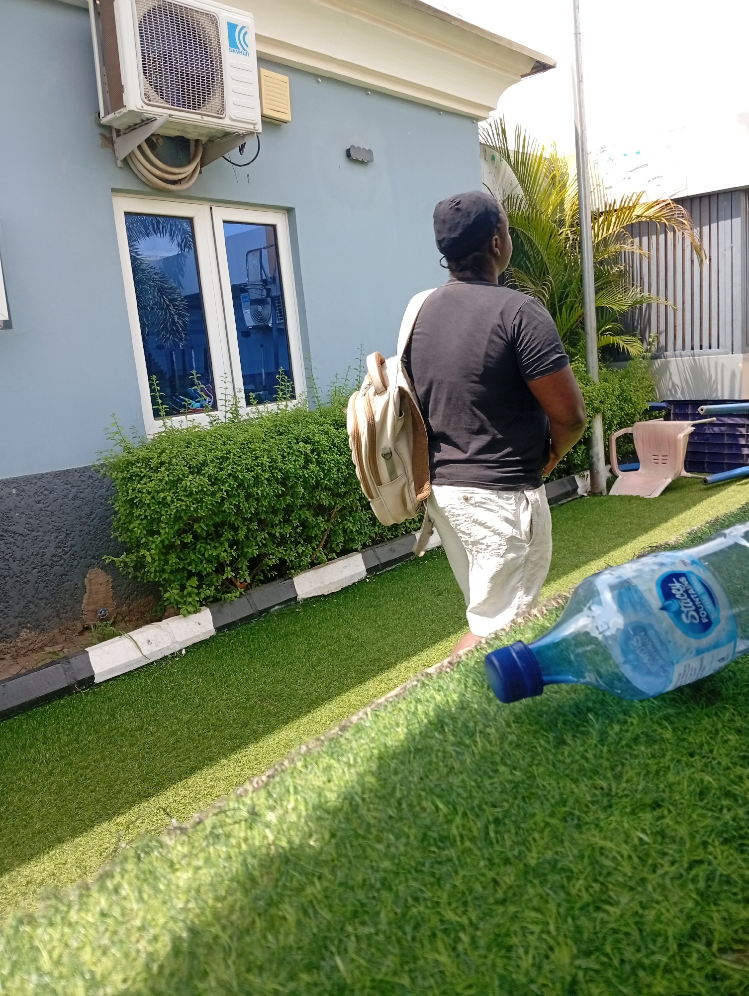 The image captures a daytime scene outdoors, possibly in a residential or commercial area of Adankolo, Nigeria. In the foreground, a discarded transparent plastic water bottle with a blue cap lies on artificial grass, partially obscuring the view. Behind it stands a person with their back to the camera, wearing a black t-shirt, light-colored shorts, and a black cap. They have a tan-colored backpack slung over their shoulder.
To the left of the person, there is a light blue building with a window reflecting foliage. An air conditioning unit is mounted on the wall above the window. In front of the building, there is a row of green bushes bordering the edge of the artificial grass, marked with black and white concrete blocks.
Behind the person, there is a small palm tree with yellowing leaves. In the background, there is a white building with metal gates. A plastic chair and a blue plastic crate are visible. There is also a metal pole rising in the background. The scene is brightly lit with sunlight casting shadows on the artificial grass.