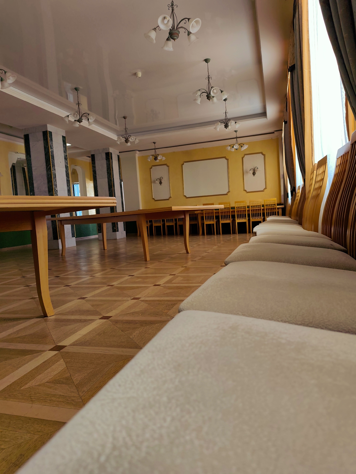 The image shows a room in what appears to be a formal meeting space, possibly in the city of Chaikovsky, Russia. In the foreground, there's a row of upholstered chairs. Behind them are wooden tables and chairs set up as if for a conference. The walls are painted a light yellow color, adorned with framed blank white rectangles and decorative wall fixtures. Marble-clad columns support the high ceiling. Above, chandeliers are affixed to the ceiling. The floor is parquet wood with a geometric pattern. Curtains partially cover a window on the right side.