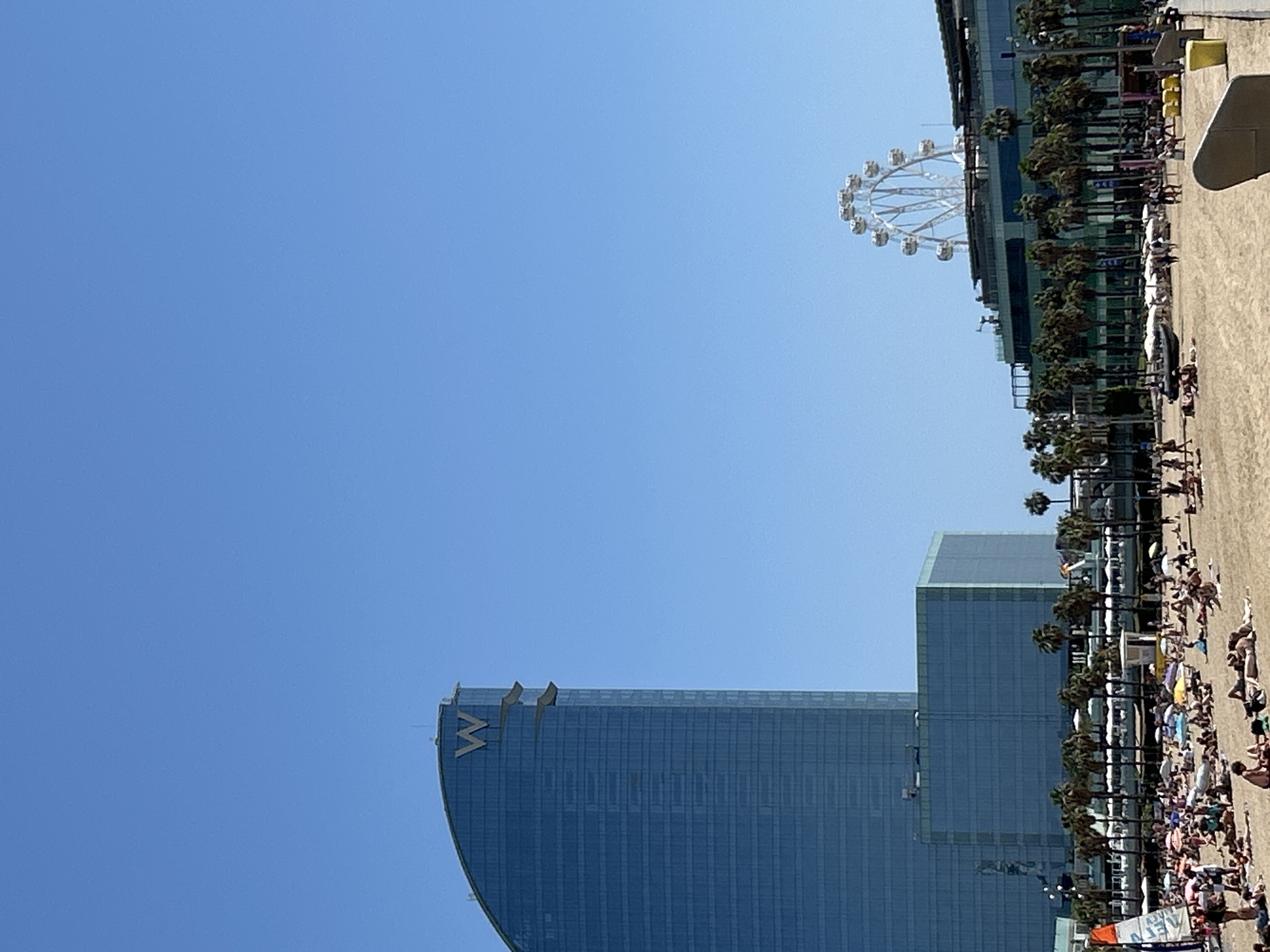The image depicts a sunny day at Barceloneta Beach in Barcelona, Spain. In the background, the distinctive sail-shaped W Hotel, a prominent landmark, stands tall. To its right, a Ferris wheel, likely the "Parc d'Atraccions Tibidabo," is visible above modern buildings and a line of palm trees. The foreground is filled with a crowded beach scene, with numerous people sunbathing, relaxing on towels, and some walking on the sand. Several umbrellas and beach chairs are scattered across the beach. A small sailboat is positioned on the left side of the frame. The sky is clear and blue, indicating fair weather.