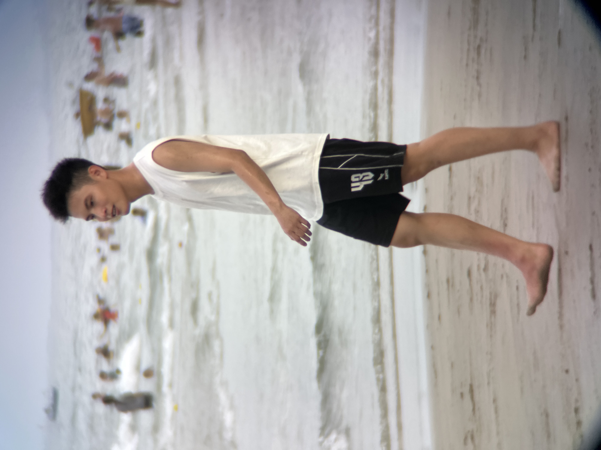 A young Asian man with dark, spiky hair walks barefoot on a sandy beach in Đà Nẵng, Vietnam. He is wearing a sleeveless white t-shirt and black athletic shorts with white trim and the number "43" printed on the left leg. The man is facing to his left and appears to be walking towards the right side of the frame. In the background, several people are visible in the ocean, some swimming and others wading. The water is choppy with small waves. A boat is present in the far distance on the left side of the image. The sky is overcast, creating a diffused light over the scene. The sand is light brown and appears damp near the water's edge.