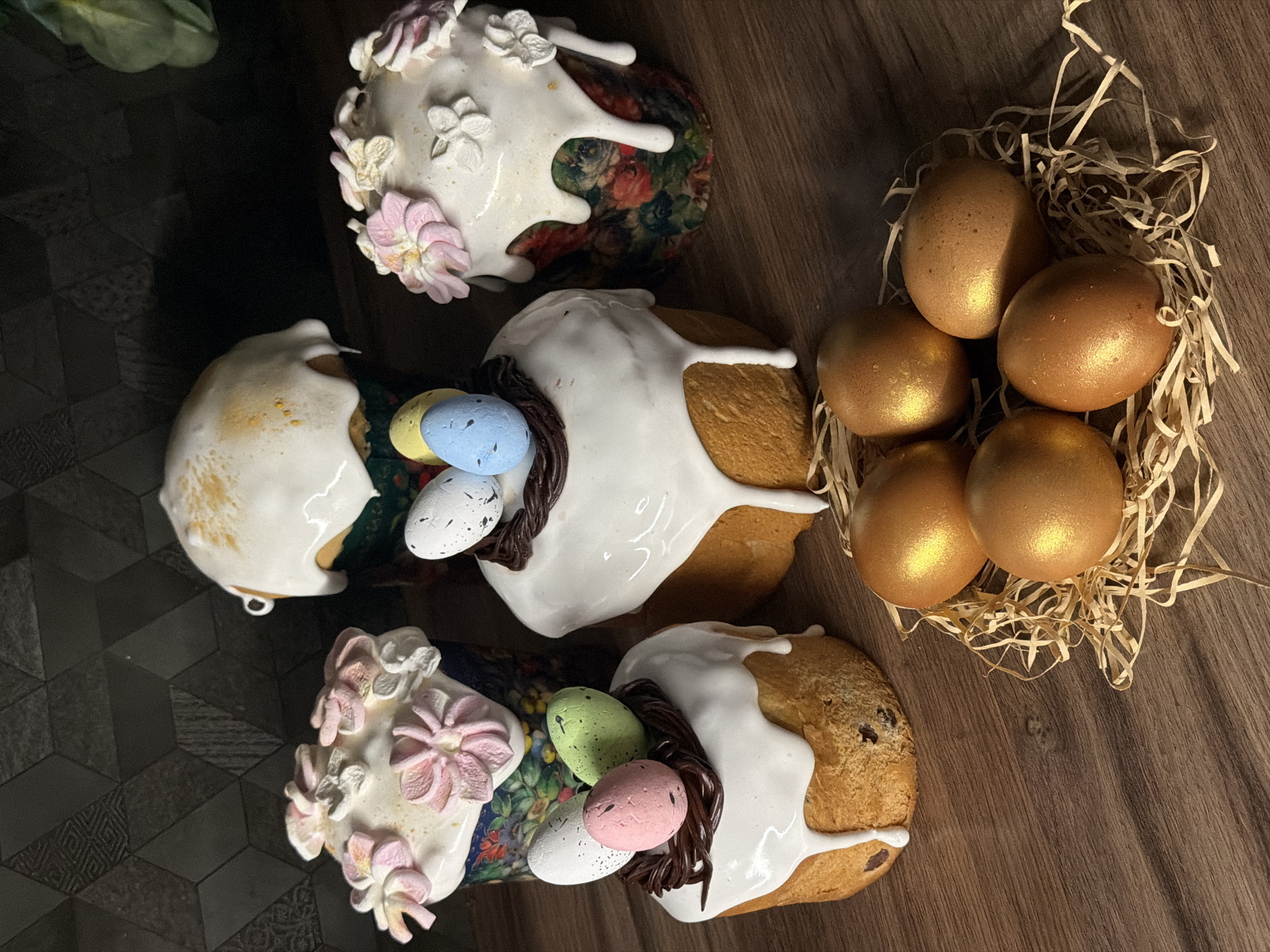 The image depicts a still-life arrangement of Easter treats, likely taken indoors. The main focus is on four Easter cakes (kulich), each decorated with white icing that drips down the sides. Two cakes have floral decorations in pastel pink and white icing. The other two cakes have nests made of dark chocolate strands, holding small, colorful speckled eggs. Next to the cakes is a nest of straw-like material containing six golden-colored Easter eggs. The cakes and eggs are set on a dark wooden surface. The location, Perm, Russia, is not discernible from the image.