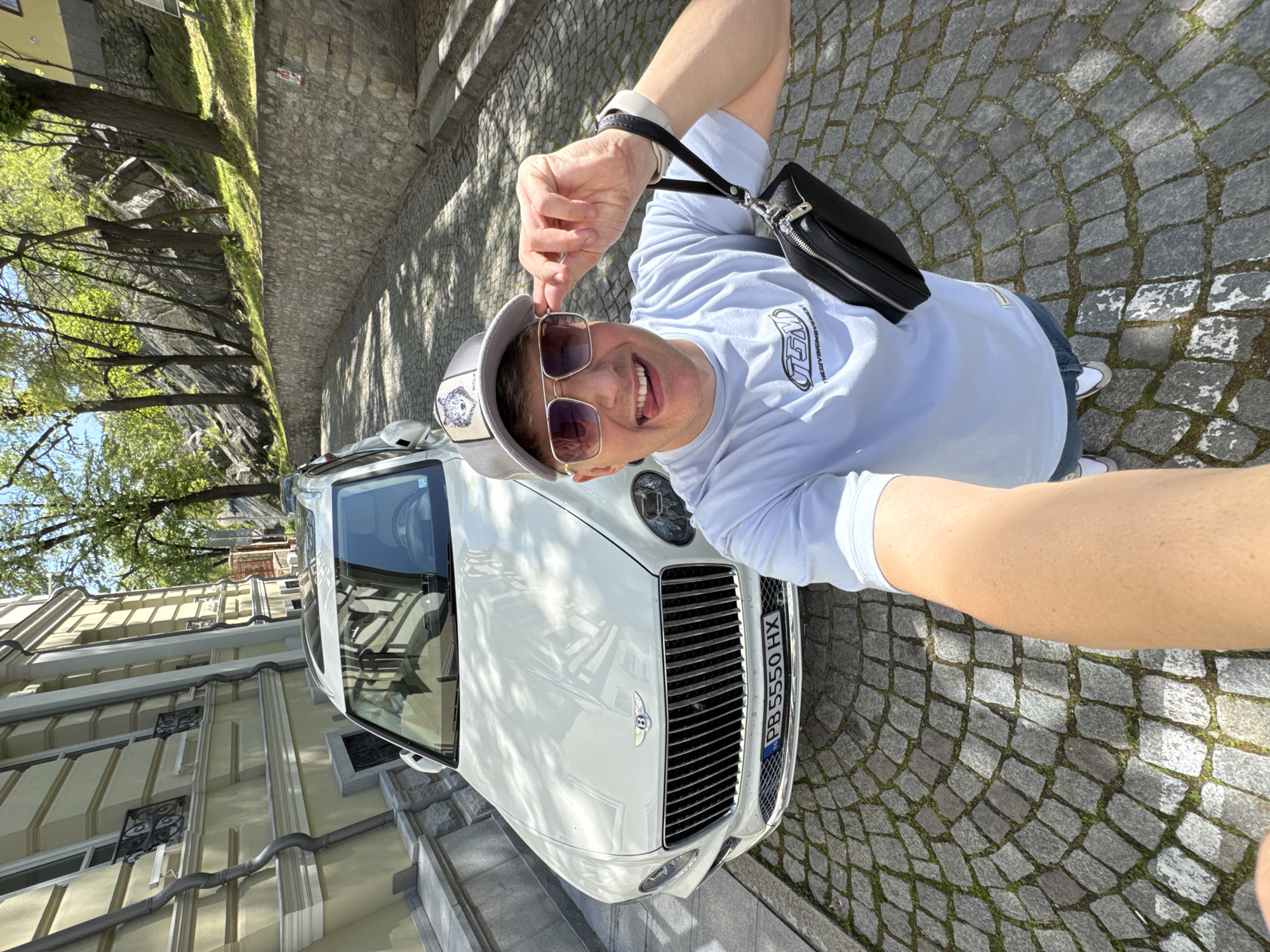 A man is posing for a selfie standing in front of a white Bentley car on a cobblestone street. The man wears a light blue t-shirt, blue jeans, white sneakers, sunglasses, and a gray cap with a wolf emblem. He's smiling and holding up his sunglasses slightly. A black crossbody bag is visible. The car has a license plate "PB 5550 HX". A building with light yellow walls and wrought-iron window grilles is visible behind the car. A tree line is also visible. The location is in the city of Plovdiv, Bulgaria.