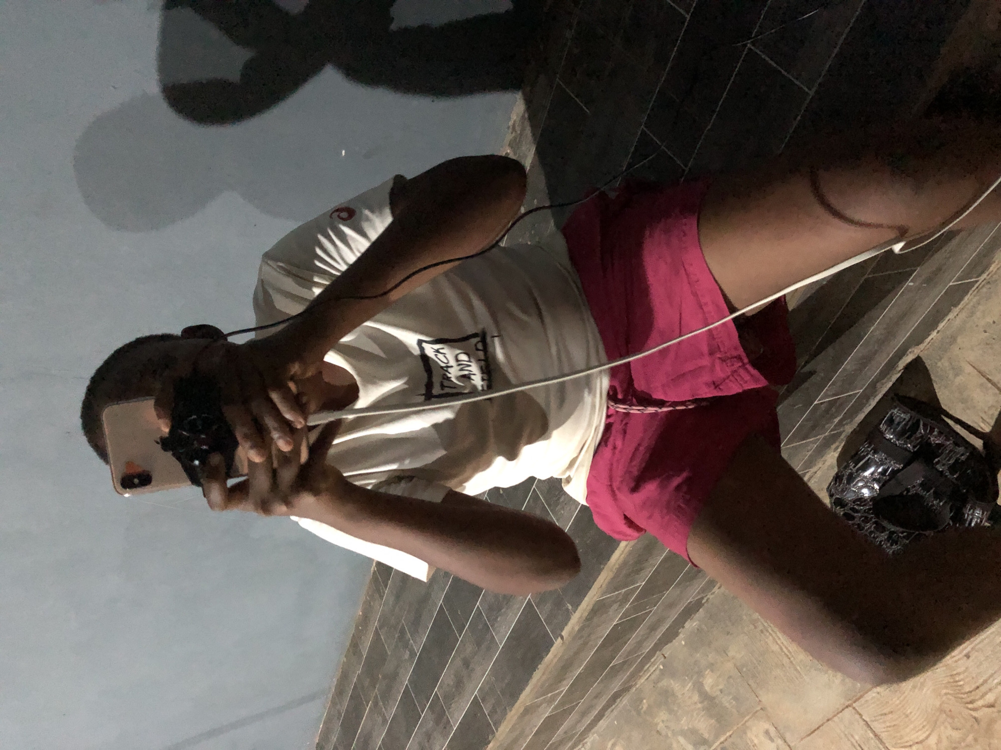 A person is using a smartphone. The person is wearing a white t-shirt with a black design and pink shorts. A black bag is on the ground next to the person. The location appears to be outdoors on a tiled surface, possibly a sidewalk. The city and state are unknown.