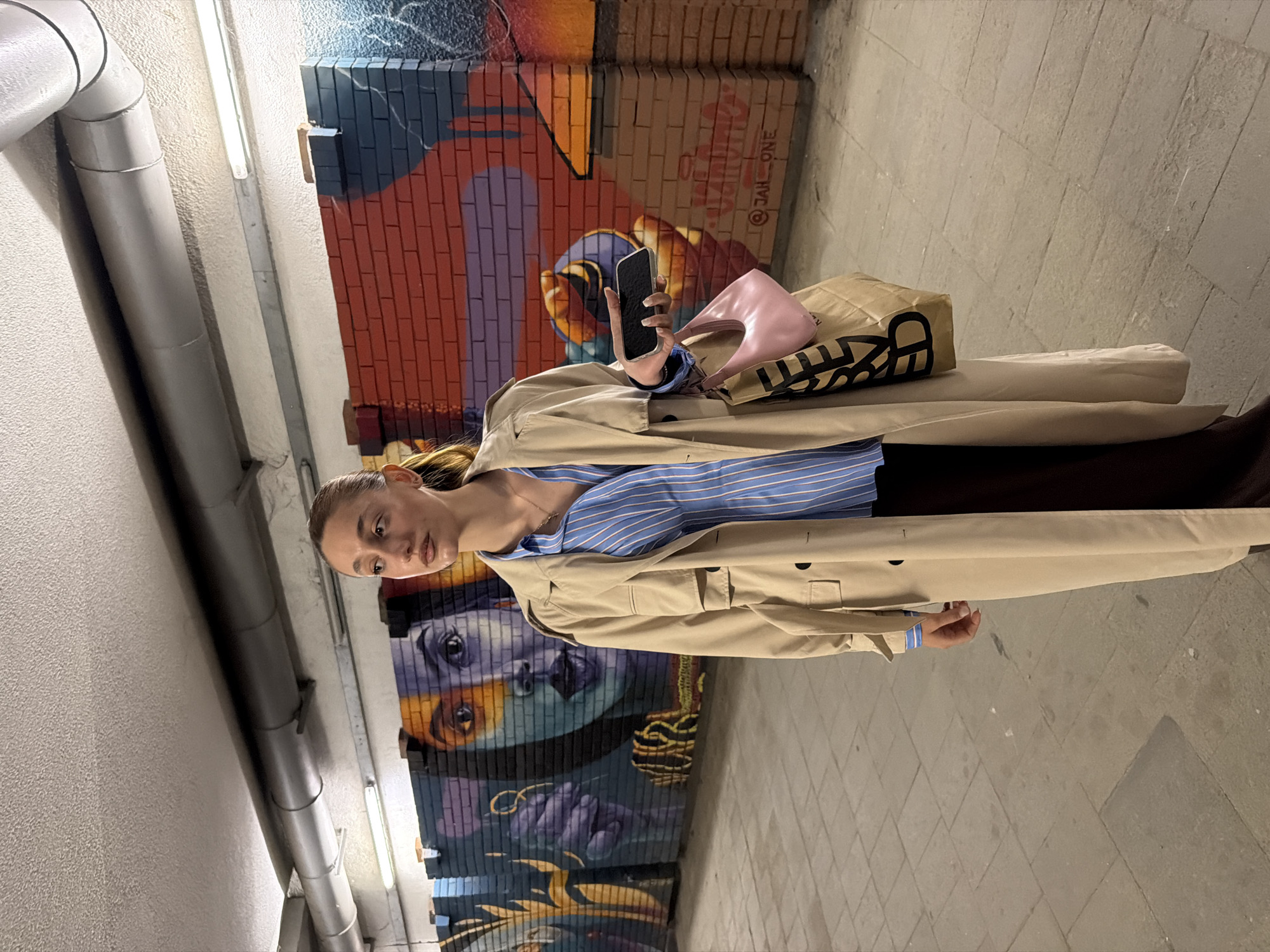 A woman stands in what appears to be a pedestrian underpass. She is holding a smartphone in her left hand and a pink handbag and brown paper bag on top of a tan trench coat. She is wearing a blue and white striped shirt. The underpass wall behind her features a mural of faces, and some graffiti. The floor is tiled. There are overhead pipes along the top of the left wall, illuminated by strip lighting. The photo was taken indoors, likely in the city of Sofia, Bulgaria.