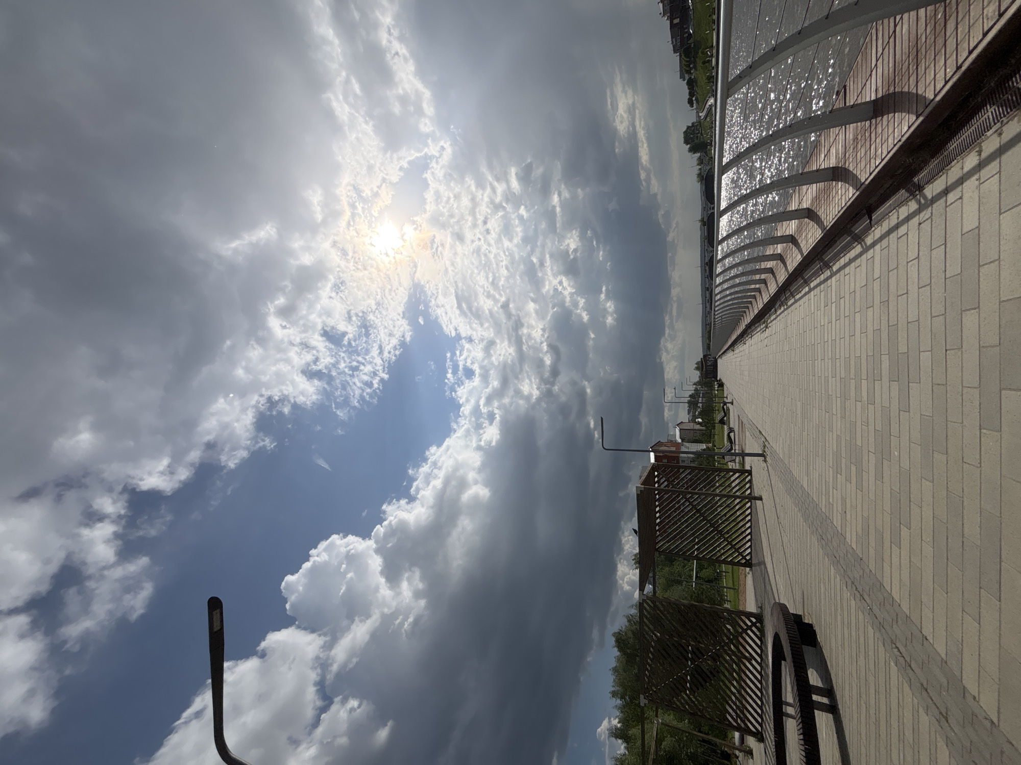 The image shows a pathway in Votkinsk, Russia, under a sky with a bright sun and heavy clouds. The pathway is constructed of light-colored rectangular pavers with a dark gray stripe running down the middle. Along the right side of the path is a metal railing with a screen behind it, reflecting sunlight. Benches with wooden lattice covers are positioned on the left side of the pathway, along with a few trees and grass. A tall, curved lamppost is visible on the far left, reaching into the sky. In the distance, buildings are visible.