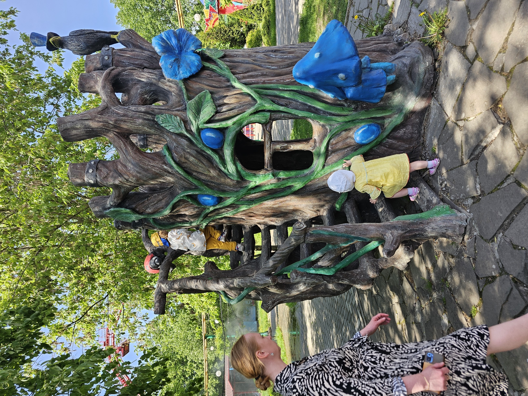 The image shows a person taking a picture of a child playing on a tree-like structure in a park. The structure is made of concrete and painted to resemble a tree, with blue flowers and mushrooms. Two children are climbing on the structure. The person taking the picture is standing on a stone path. The location is Khimki, Russia.