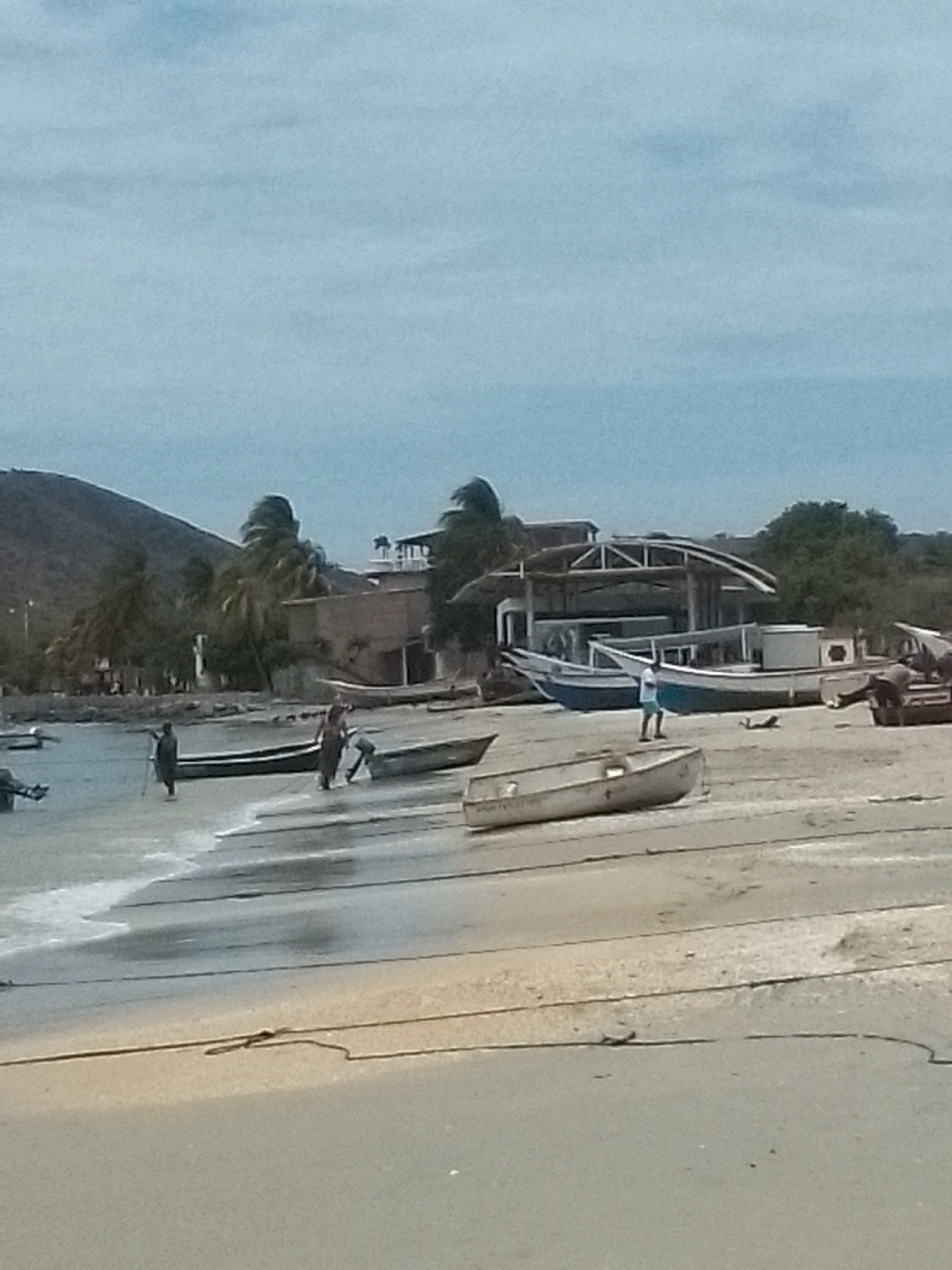 The image shows a beach scene in Juan Griego, Venezuela. Several fishing boats are pulled onto the sand. A few people are wading in the shallow water near the boats. In the background, there are buildings and some palm trees, with a hillside visible on the left. The sky is overcast. The scene is composed of sand, water, boats, people, buildings, and natural elements.