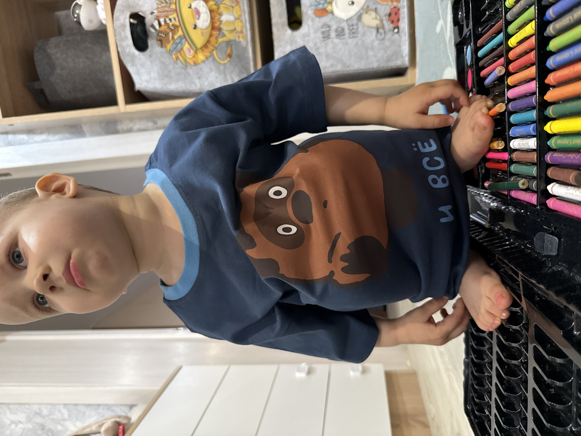 A young child with light skin and hair is depicted in an indoor setting, possibly a home in Rusanovo, Russia. The child is wearing a navy-blue long-sleeved shirt with a cartoon bear print. The child's hand and foot are positioned near a box of colorful oil pastels. The backdrop features storage bins with animal-themed designs. The environment includes a white radiator and what appears to be a wooden cabinet or shelf.