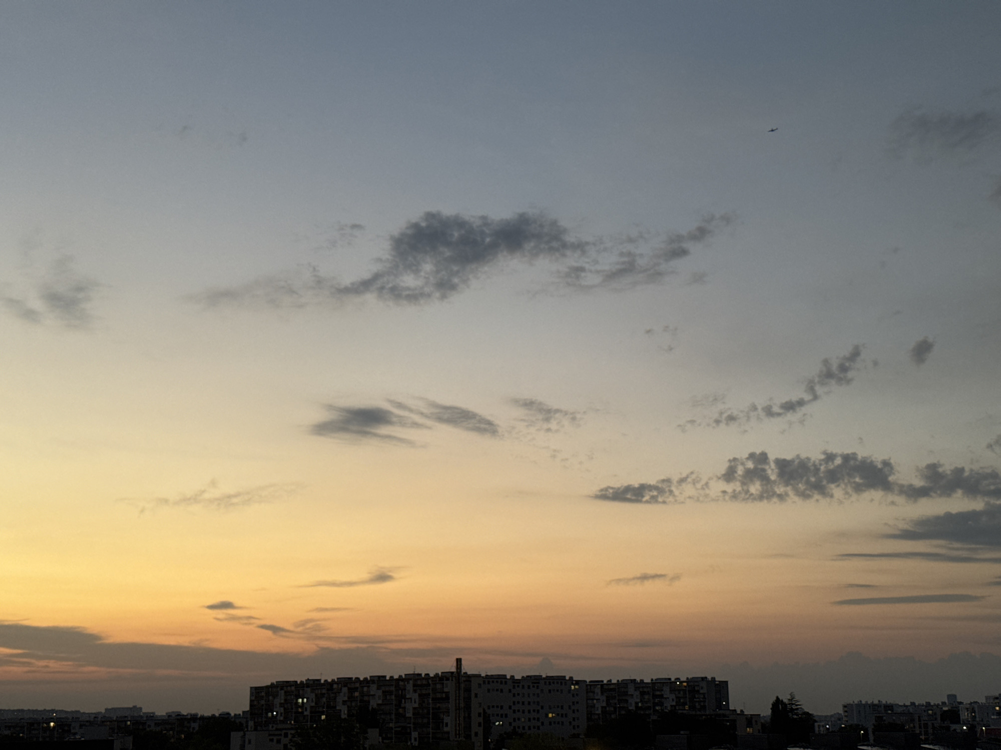 This is a horizontal, eye-level, medium shot of a sunset over a city skyline. The sky dominates the frame, transitioning from a pale blue at the top to a warm yellow and orange near the horizon. Wispy, dark clouds are scattered across the sky, with a tiny airplane visible in the upper right quadrant. Below the sky, a dense city skyline stretches horizontally across the bottom of the image, featuring numerous apartment buildings. The buildings are dark silhouettes against the colorful sky, with some windows appearing lit. The overall mood is peaceful and serene, suggesting the end of a day. The time of day is clearly sunset, indicated by the gradient of colors in the sky. No text is visible in the image.