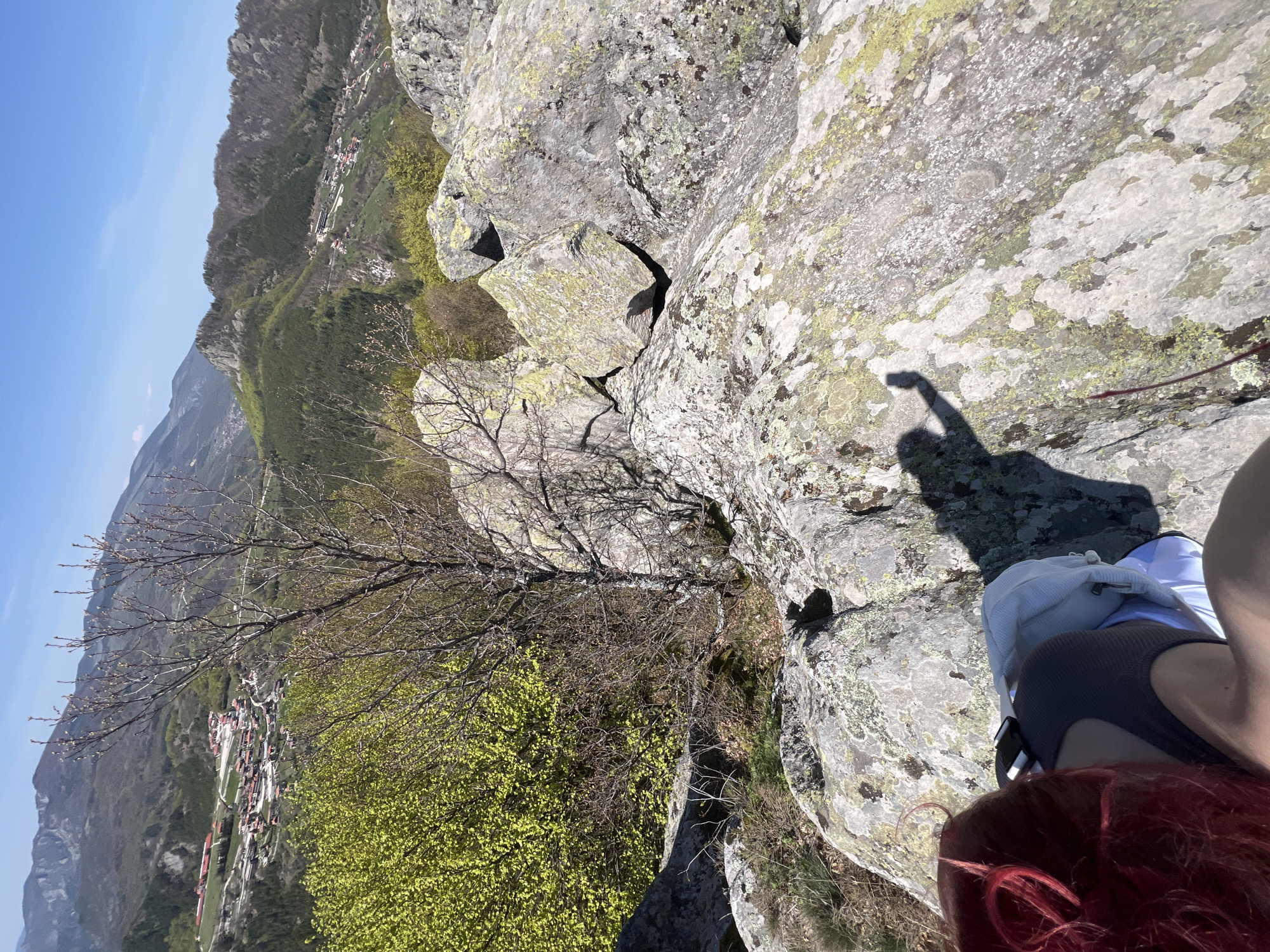 The image captures a scenic view from a rocky mountain peak or outcrop, looking down into a valley. The perspective is taken from a high vantage point, with the bottom right showing the top of a person's head with bright red hair, wearing a dark top, and a white bag or jacket beside them. A prominent shadow of a person's hand, appearing to be holding a phone, is cast on the sunlit, grey rocks in the immediate foreground. These rocks are extensively covered in patches of green and yellow lichen.
Below the immediate rocky ledge, the terrain drops steeply, revealing a mix of trees. Some trees have delicate, bare branches, while others are adorned with fresh, vibrant light-green foliage, indicative of early spring. Further in the midground, nestled within the valley and surrounded by forested hills, is a small village or town with numerous buildings, some featuring reddish roofs, stretching along the landscape.
The background consists of successive layers of mountains and hills, densely covered in trees, receding into the distance. The sky above is clear, expansive, and a brilliant blue, suggesting a sunny day. The strong sunlight casts distinct shadows, indicating that the photo was taken during the late morning or early afternoon. The overall setting points to a mountainous, natural environment, specifically Sini Vrah, Bulgaria, characterized by rugged beauty and a panoramic vista. No visible text is present in the image.