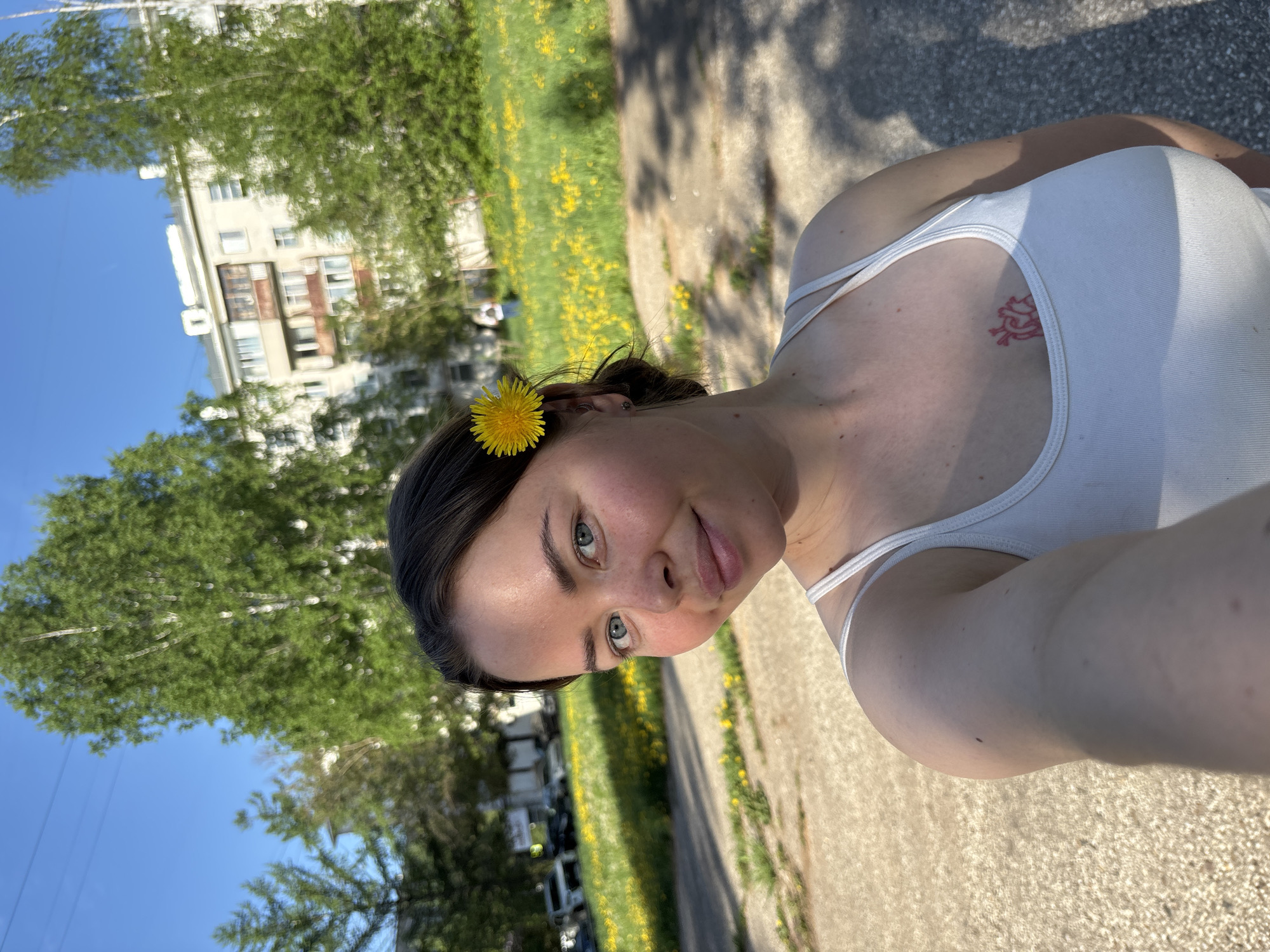 A young woman in a white tank top takes a selfie outdoors in Izhevsk, Russia. She has a yellow dandelion tucked behind her ear. A red tattoo is visible on her chest. In the background, there is a grassy area dotted with yellow dandelions, a sidewalk, trees, and a light-colored building. The sky is blue.