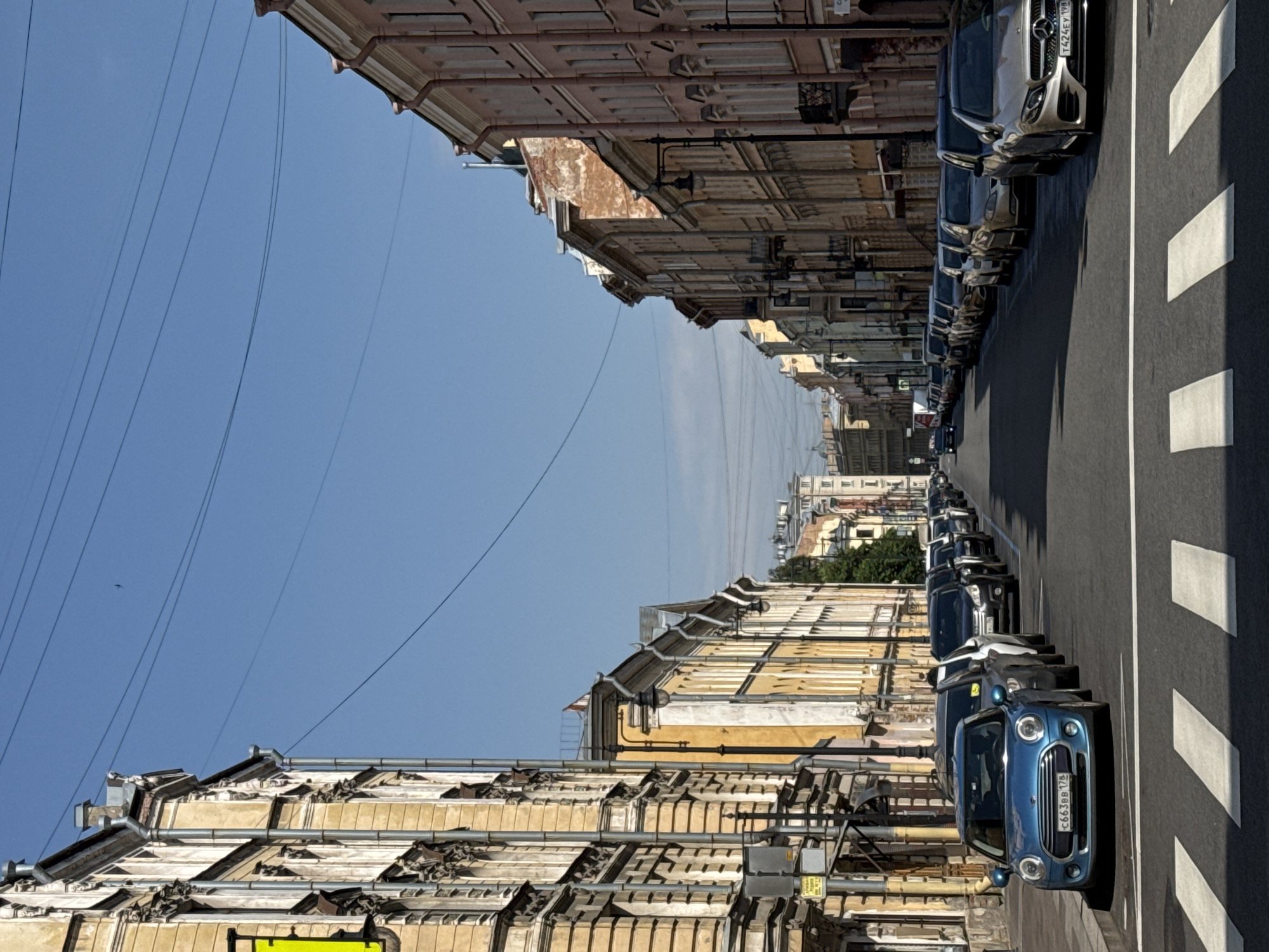 This is a full-shot, eye-level, vertical image of a street in Saint Petersburg, Russia, on a sunny day. The street is lined with multi-story buildings on both sides, featuring architectural details such as ornate facades, balconies, and mansard roofs. The buildings are painted in various warm tones, including yellow, beige, and pink.
A row of parked cars is visible along the right side of the street. The closest car in the foreground is a silver Mercedes-Benz. A pedestrian crossing with white stripes is painted on the asphalt in the lower foreground. The sky is clear and blue with some thin, dark power lines crisscrossing overhead. A single bird is visible as a small speck in the sky above the buildings on the left. The overall impression is of a calm, urban street scene.