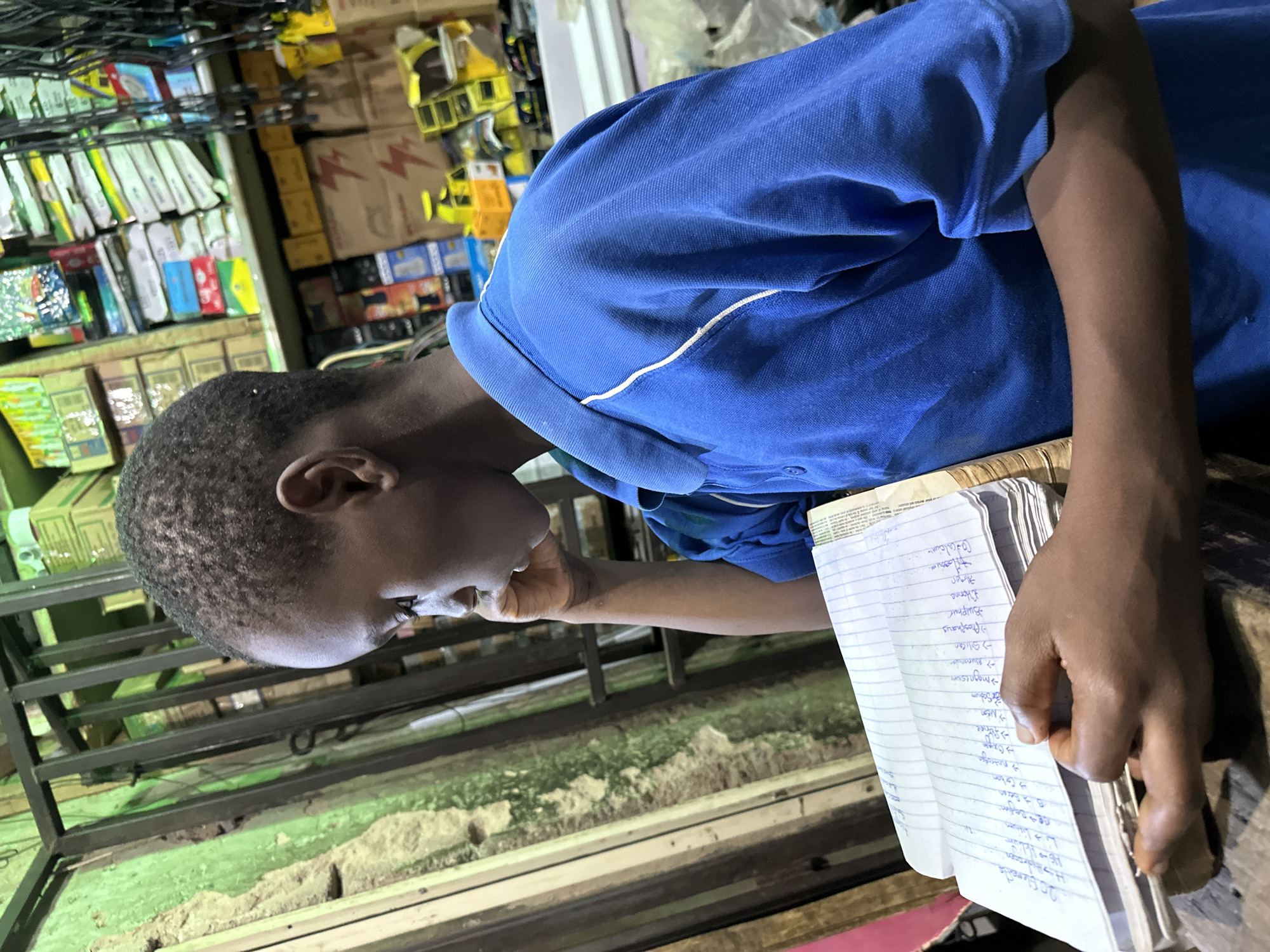 A young boy, likely of Nigerian descent, is seated indoors, holding an open notebook. The notebook is filled with handwritten notes. The boy is wearing a blue polo shirt. He is leaning his head against his hand, appearing to be concentrating on the notebook. The background suggests a cluttered environment, possibly a market stall or workshop, with shelves filled with various goods, boxes, and electrical wires. The lighting is somewhat dim, casting shadows and highlighting certain objects. The location is identified as Narayi, Nigeria.