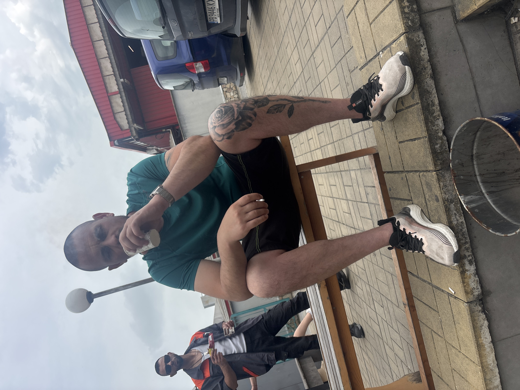 The image, oriented vertically and rotated 90 degrees counter-clockwise, captures a low-angle outdoor scene featuring two men under an overcast sky.
The primary subject, a man with a shaved head, is seated on a wooden bench, looking directly at the camera with a neutral expression. He wears a dark green t-shirt, black shorts, and light-colored athletic shoes with black laces and pink accents. A prominent black and grey rose tattoo covers his left upper leg. He has a silver-toned watch on his left wrist and holds a small white paper cup to his mouth.
Behind him, another man is partially visible in the background, wearing sunglasses, a white t-shirt, and a dark work-style jacket with orange accents. He appears to be holding a bottle or can and possibly a cigarette.
The ground is paved with light-colored rectangular tiles. In the background, a building with a red corrugated roof is visible, along with parked cars. A dark grey/black car displays a license plate "PB 9446 KX", and a blue car has "DACIA" partially visible on its rear. A street lamp with a white spherical light stands behind the main subject's head. On the right side of the frame, near the main subject's foot, is a blue metal bucket with the partial text "ITAL". A discarded cigarette butt is visible on the pavement. The scene appears to be a casual daytime gathering or break in Plovdiv, Bulgaria.
