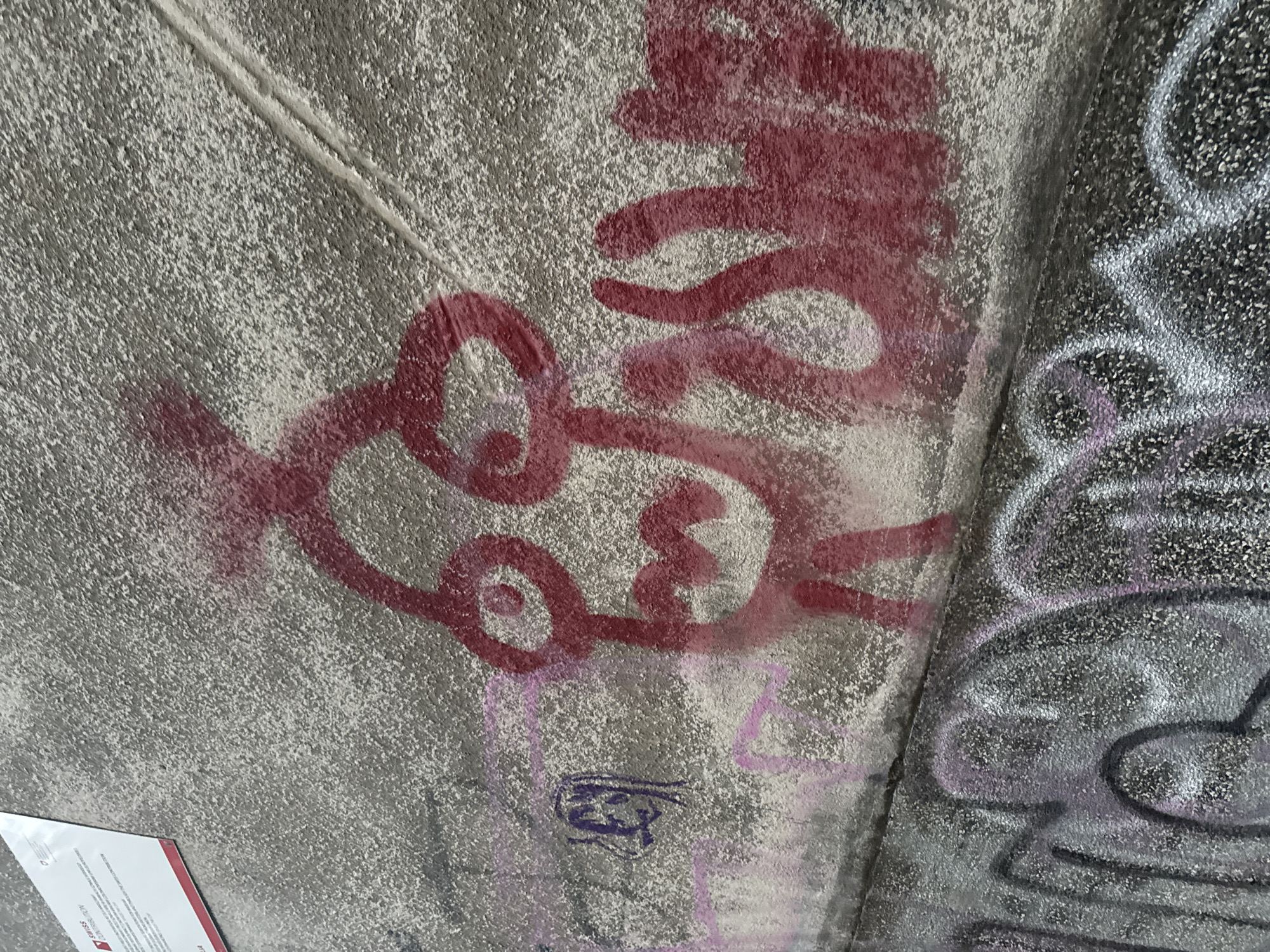 The image shows a rough concrete wall with graffiti. The main subject is a large, stylized red drawing of a face with exaggerated eyes and a wide mouth. To the left of the face, there are faint purple markings and pink streaks, suggesting layering of graffiti. The lower part of the wall is covered in more graffiti, primarily in black and silver with purple accents, forming abstract shapes and letters. A white sign with red trim and text is visible in the upper left corner of the image. The texture of the concrete is coarse and speckled.