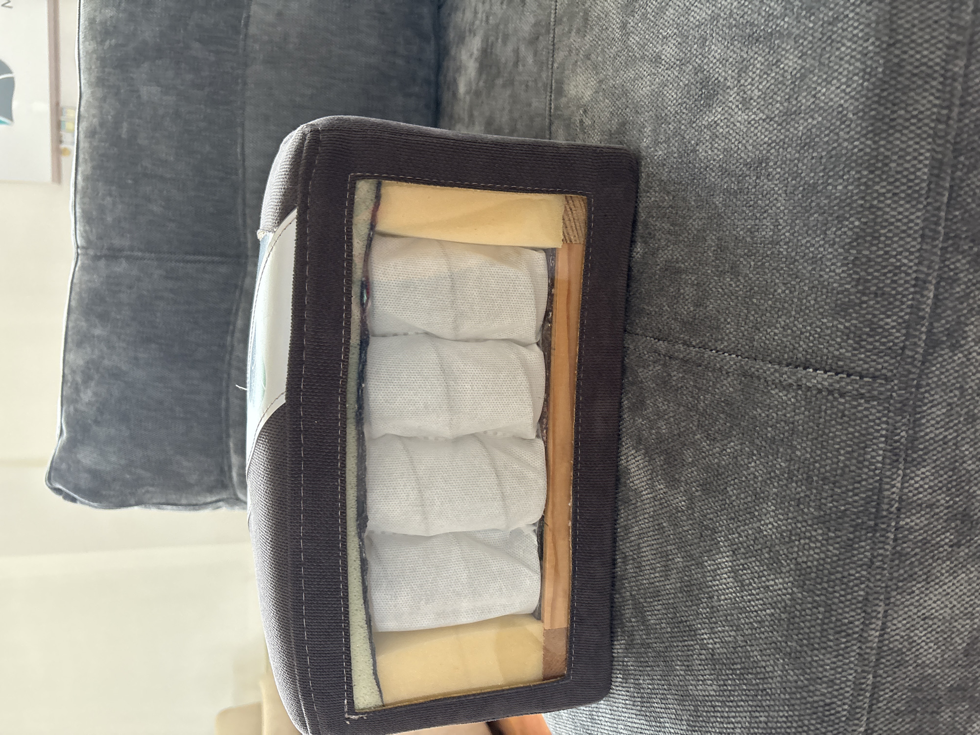 The image shows a cross-section of a sofa cushion with visible internal components. The cushion is covered in dark brown fabric and has a rectangular opening revealing its construction. Inside, there are several white, individually wrapped springs, some light yellow foam padding, and a wooden frame. The sofa itself is upholstered in a dark gray textured fabric, with a visible seam on the cushion's surface. The background suggests an indoor setting with a soft, diffused light. There is no indication of the location being in Zaporizhzhia, Ukraine.