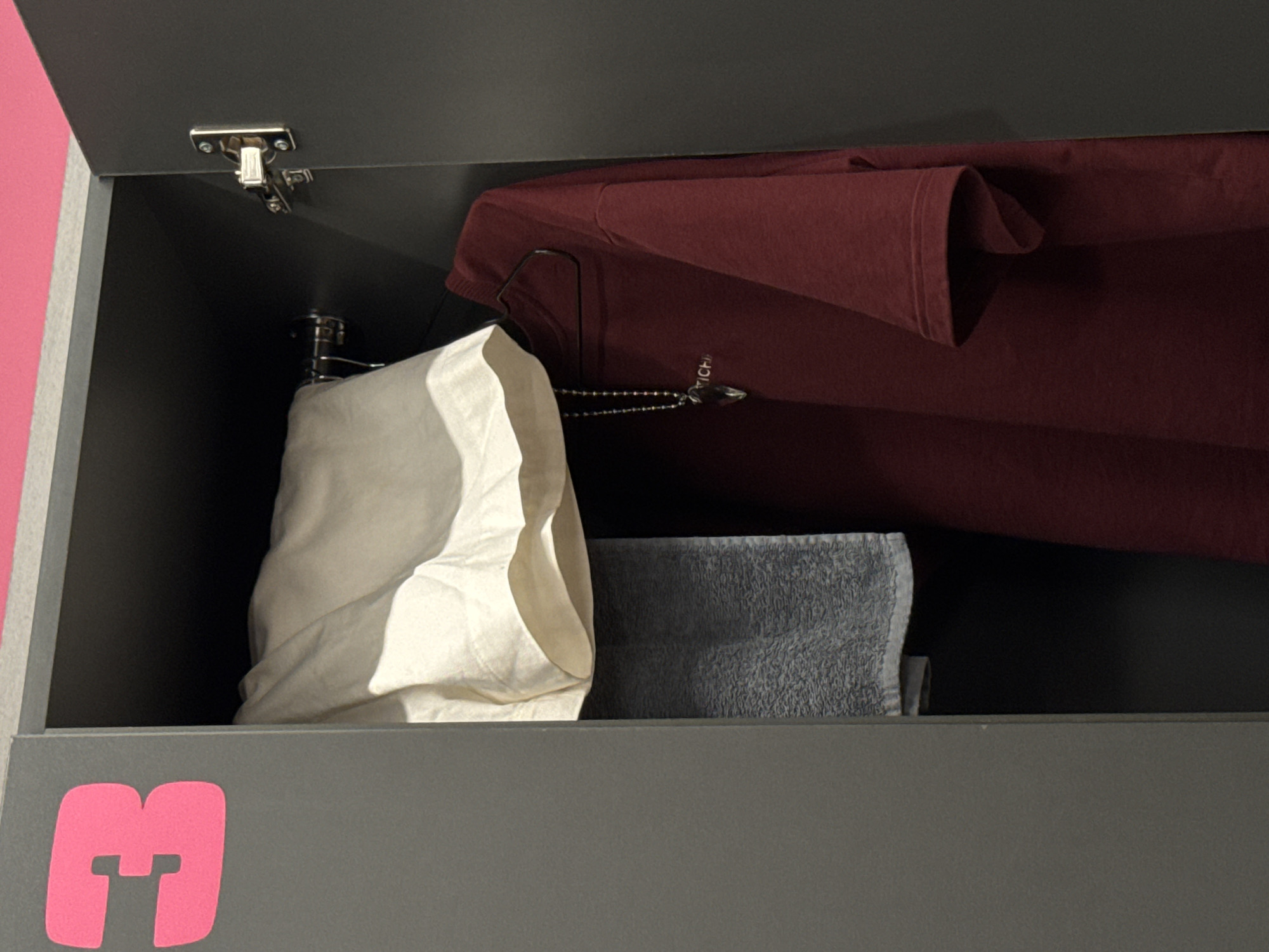 The image shows a close-up view of an open grey locker. On the left side of the locker door, a large, bright pink number "3" is visible. Inside the locker, a light-colored garment, possibly shorts or a skirt, is draped over a hanger. Below this, a grey towel is folded and hanging. To the right of the towel, a maroon t-shirt is hanging on a black hanger. A silver necklace with a pendant hangs in front of the t-shirt. The locker appears to be in a public facility, such as a gym or swimming pool, given its design and the presence of a numbered locker. The location is identified as Sochi, Russia.