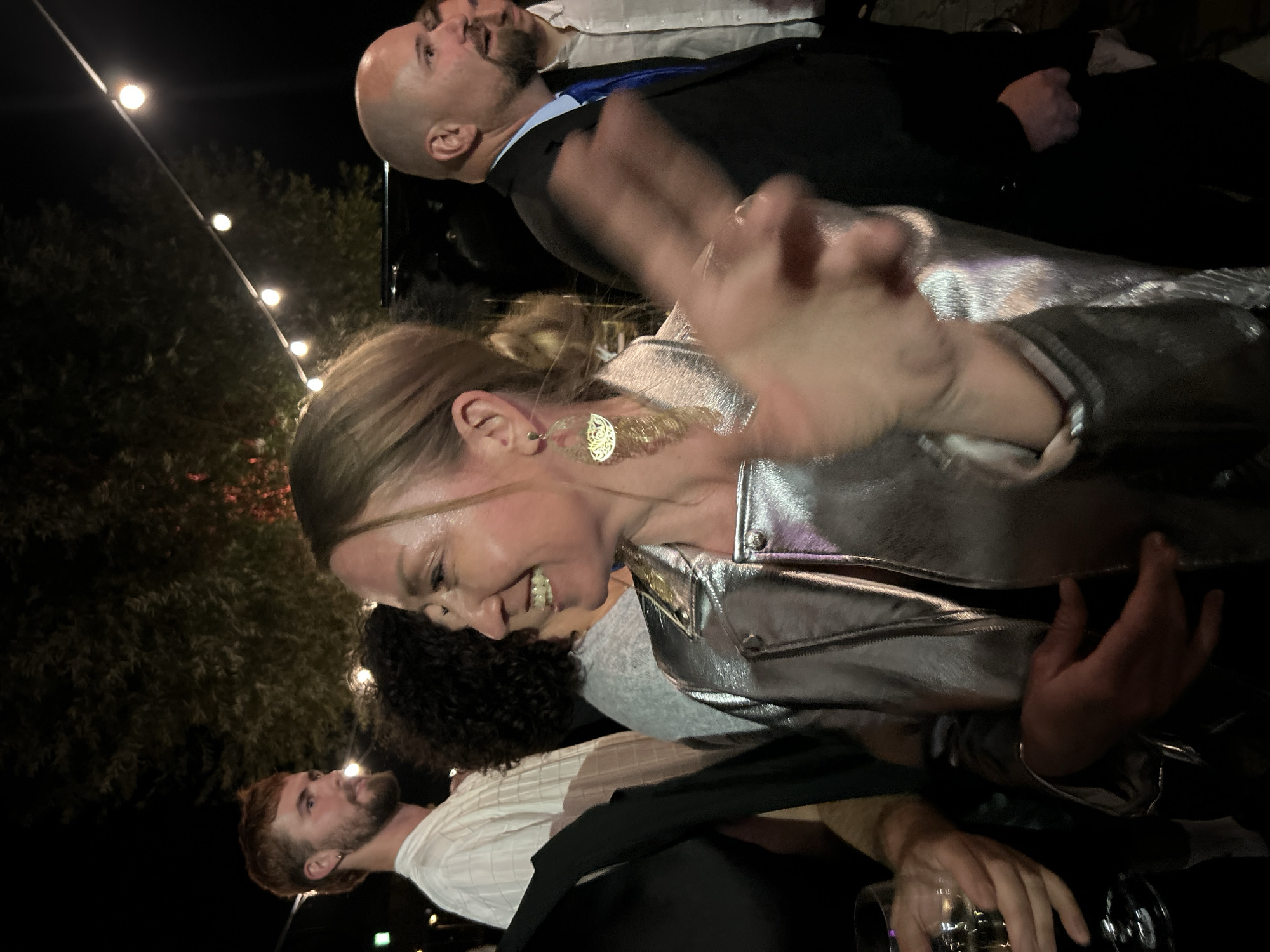 The image depicts an outdoor nighttime gathering, likely in Berlin, Germany, illuminated by string lights. In the foreground, a woman with blonde hair pulled back smiles and gestures towards the camera. She wears a shiny silver jacket and large, ornate gold earrings. To her left, a person holding a glass of white wine is partially visible. To her right, a bald man in a suit and blue tie is looking away from the camera. Behind the woman, a man with reddish-brown hair and a beard, wearing a white collared shirt, is also visible in profile. The background features trees and the dark night sky, with the string lights creating bokeh effects. The overall atmosphere suggests a casual social event or party.