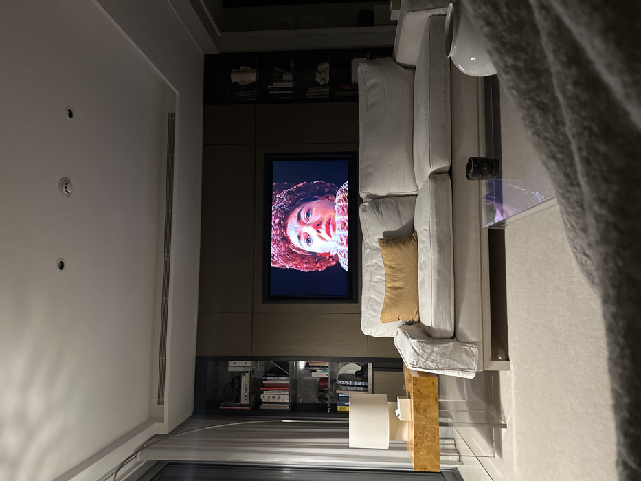 A well-lit living room displays a large TV set into a wooden built-in shelf unit, showing a woman's face. A white sectional sofa with a tan throw pillow sits adjacent to the unit. A glass-topped coffee table and a wooden side table are placed near the sofa. The room has built-in bookshelves and a window providing ample light. The overall color palette is neutral, with whites, tans, and browns dominating the scene.