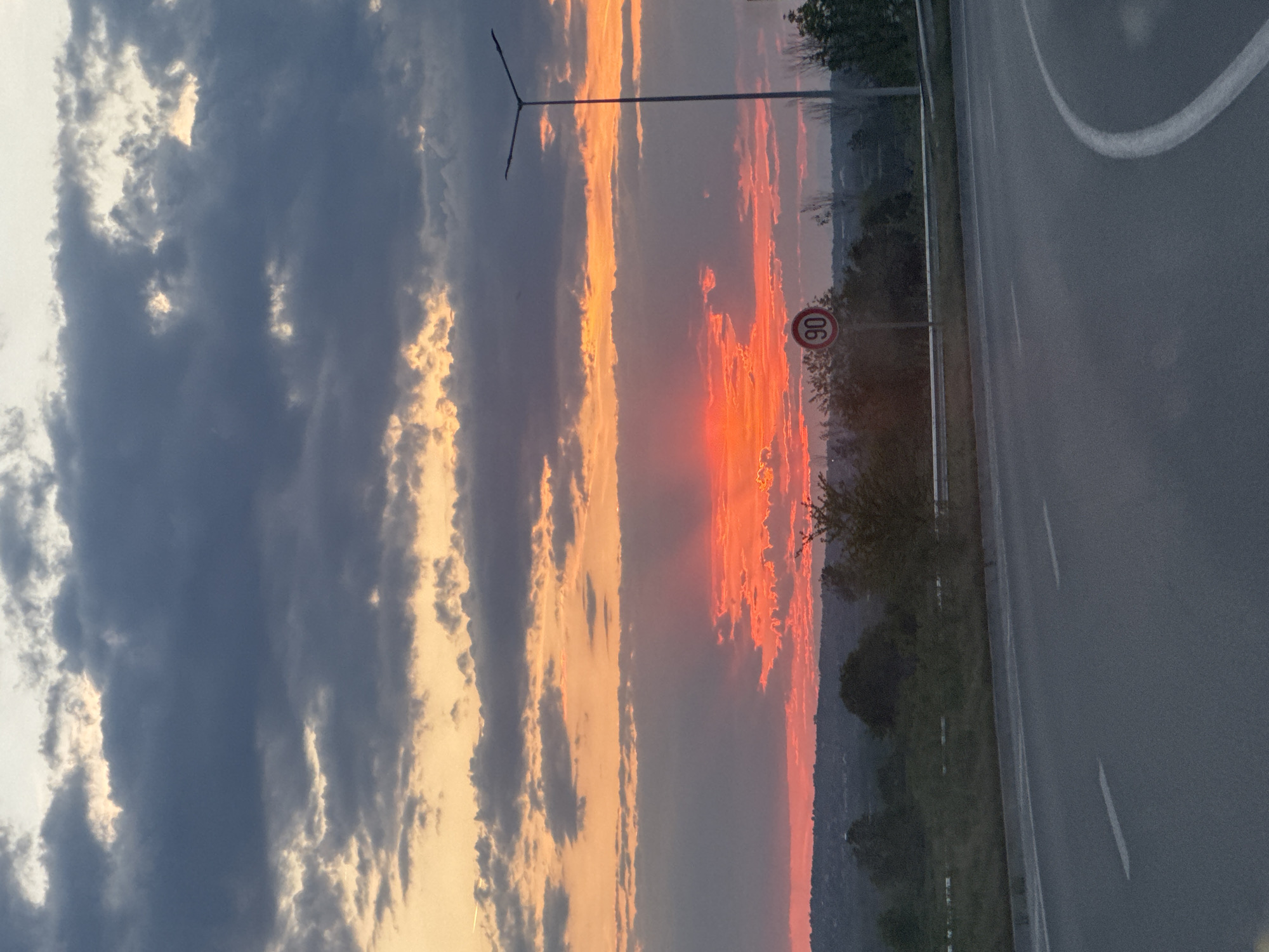 The image depicts a sunset along a roadside in or near Asenovgrad, Bulgaria. The sky displays a gradient of colors, ranging from dark, partly cloudy regions on the left to fiery orange and red hues towards the right, indicating the sun's position near the horizon. A tall street lamp stands near the edge of the road. The road itself is asphalt and features painted lane markers. A traffic sign displaying "90" within a red circle, signifying a speed limit of 90 kilometers per hour, is visible along the edge of the road. Some vegetation, including trees and bushes, lines the roadside.