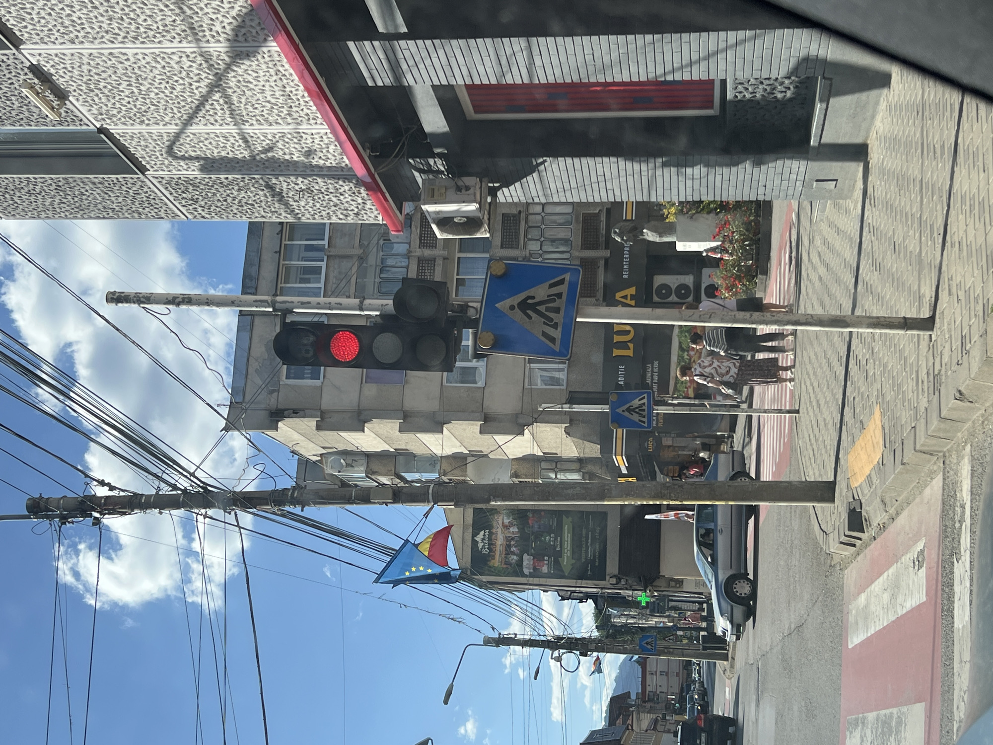 The image depicts a street scene in Câmpulung Moldovenesc, Romania, on a sunny day with scattered clouds. A traffic light displays a red signal, located near a pedestrian crossing sign with a blue background and white pedestrian silhouette. Several people are visible, including a woman with a patterned dress and a woman in a striped shirt and dark pants walking on the sidewalk. A silver car is stopped at the intersection, and other vehicles are present in the background. The buildings lining the street are multi-story structures, with one featuring a rough textured facade. Overhead, a complex network of electrical wires is suspended between poles. Two flags are visible on a pole attached to one of the buildings, one appearing to be the European Union flag and the other the Romanian flag. A billboard with text in Romanian is also displayed on the side of a building. The ground is paved with cobblestones and features a marked pedestrian crossing with red and white stripes.
