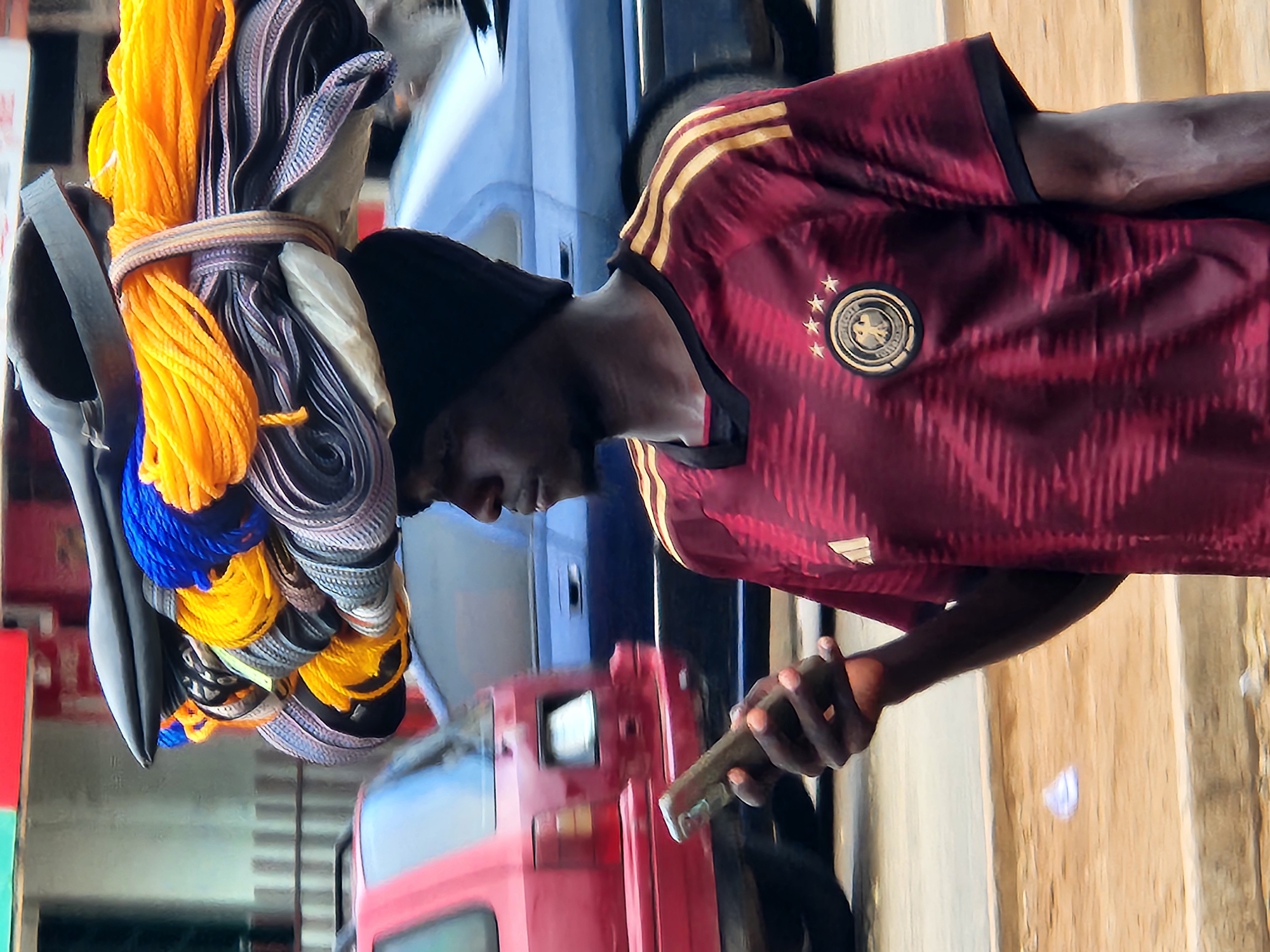 A man wearing a dark beanie and a maroon and gold striped soccer jersey, possibly representing Germany based on the crest on his shirt, walks down a street in Jos, Nigeria. He is carrying a large bundle of goods balanced on his head. The bundle includes several rolled-up items, likely textiles or mats, and a substantial amount of bright yellow and blue rope. He is holding a smartphone in his right hand, looking down at it as he walks. In the background, a red vehicle is visible to the left, and a blue sedan or SUV is behind him. The street appears to be paved, with a dirt shoulder and a concrete curb. The overall environment suggests a busy urban setting.