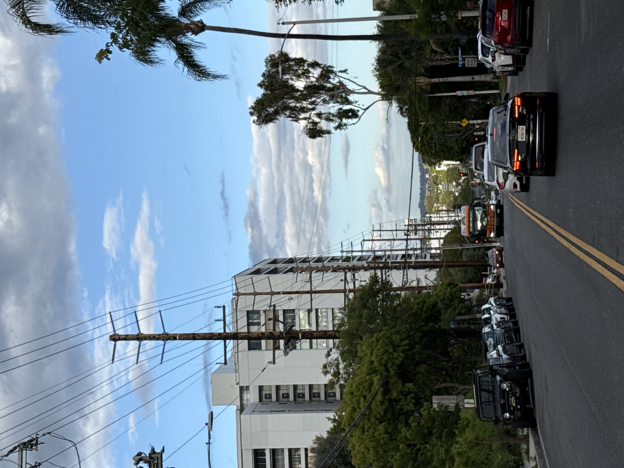 The image depicts a street scene in West Hollywood, California, USA. The view is down a paved street with double yellow lines. Parked cars are lined along the right side of the street. Moving traffic is visible in the street's lane. Buildings and utility poles line the left side of the street. Trees are visible along both sides of the street. The sky is partly cloudy. The image appears to be taken during the late afternoon or early evening.