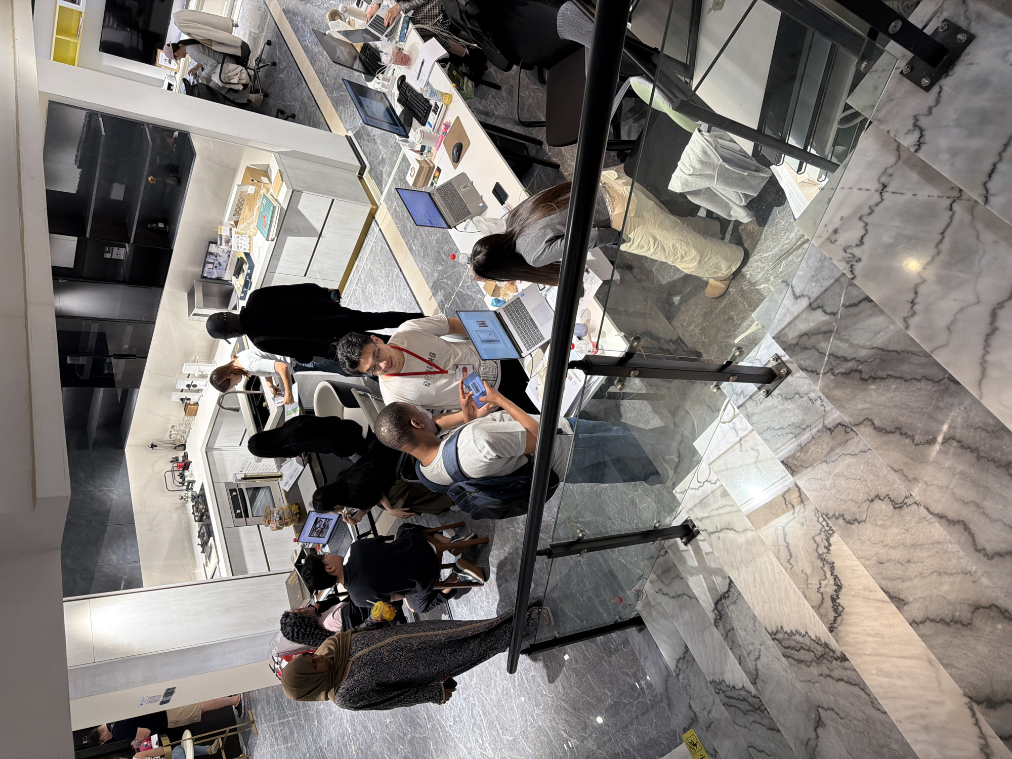 The image depicts an indoor scene, likely a modern office or co-working space, observed from a high vantage point, perhaps a mezzanine or upper floor. The location is specified as Chancheng District, China.
Several individuals are present, engaged in various activities. Some are working at desks equipped with computers and other office supplies. One person is looking at a phone. Some are interacting with each other.
The environment includes office furniture such as desks, chairs, and storage units. There's also what appears to be a kitchen area with appliances and counters. The floor is tiled with a patterned design. There are railings and glass partitions suggesting a multi-level structure. Artificial lighting illuminates the space.
The interaction and actions involve work related activities, looking at a phone and general office interactions.