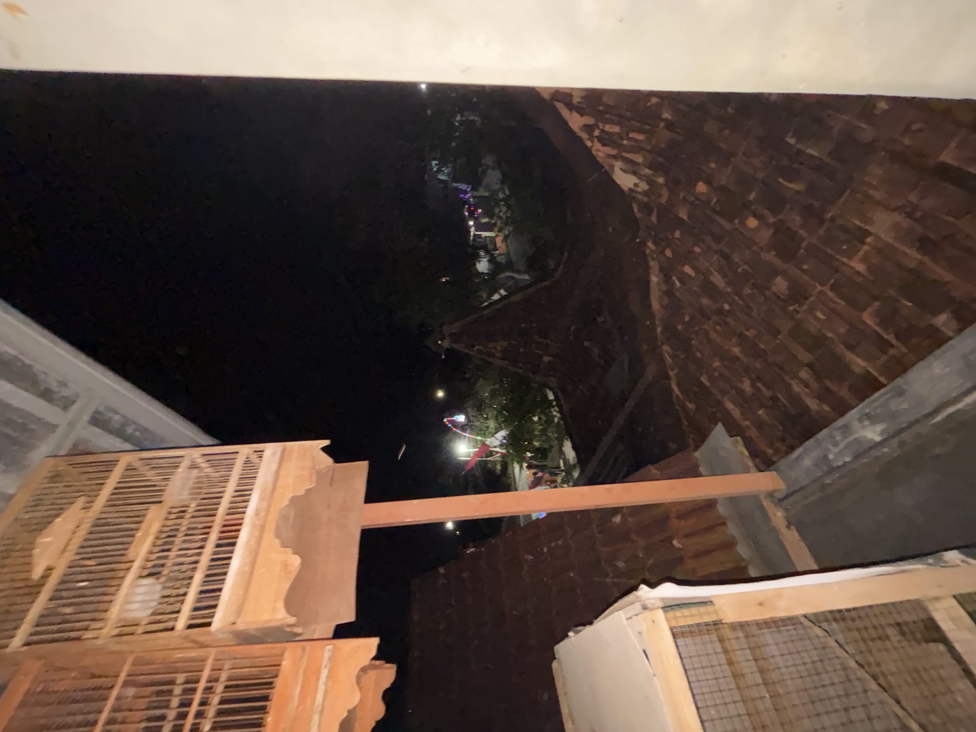 The image shows a night scene in Tirtomoyo, Indonesia, viewed from a slightly elevated position looking down onto tiled rooftops. In the foreground, there are two wooden bird cages, one partially visible on the left and a larger one in the center, supported by a reddish-brown pole. The bird cages appear to be empty.
Beyond the rooftops, a dimly lit outdoor area is visible with trees and some structures. Colorful lights are strung up, suggesting some form of celebration or market activity. A few indistinct figures can be seen in the distance, but they are too blurry to identify. The overall atmosphere is dark, with the only light sources coming from the distant decorative lights and possibly some faint illumination from houses. The architecture of the rooftops suggests a traditional style common in the region.