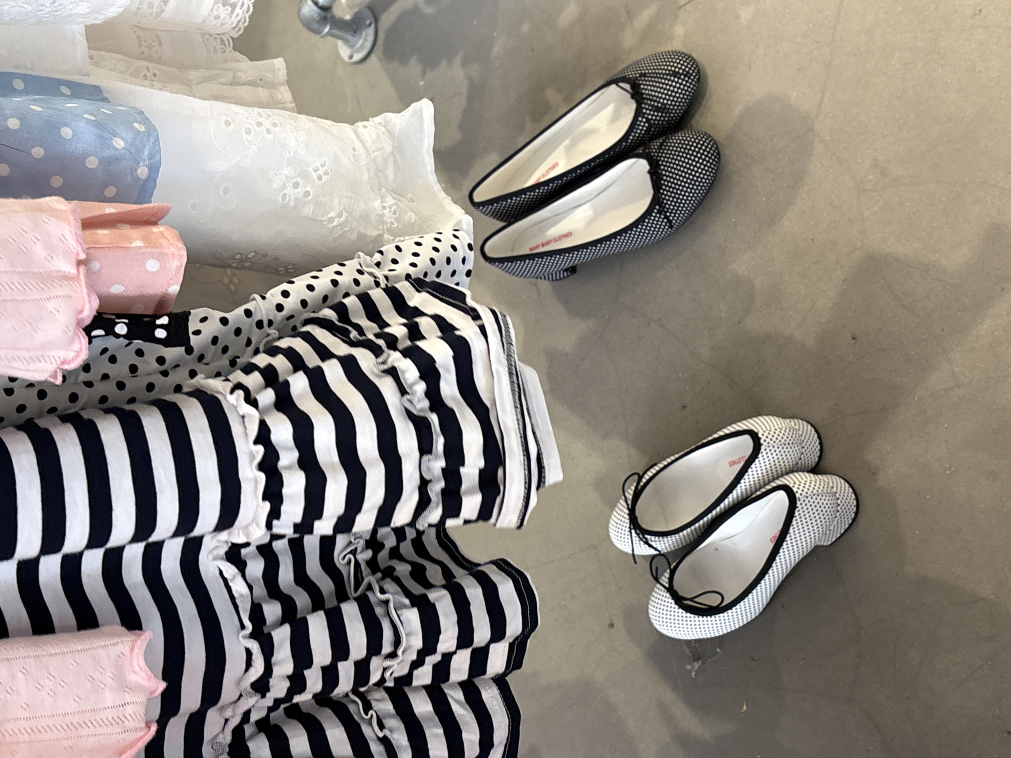 A high-angle shot shows an assortment of clothing next to two pairs of ballet flats on a grey concrete floor.
On the left side of the image, a stack of clothing dominates. At the bottom is pink fabric with a small, patterned design. Above this is horizontally striped black-and-white fabric with a ruffled edge. Layered above are fabrics in various colors and patterns including white lace, blue fabric with white dots, and pink fabric with white dots.
To the right are two pairs of ballet flats. The top pair is black with a small, white dotted pattern, while the bottom pair is white with a small, black dotted pattern.
The image appears to have been taken indoors. The presence of clothing, and ballet flats, suggest the location is likely a store, possibly in London, United Kingdom. However, there are no specific elements to confirm its location in London. There are no people visible in the shot.