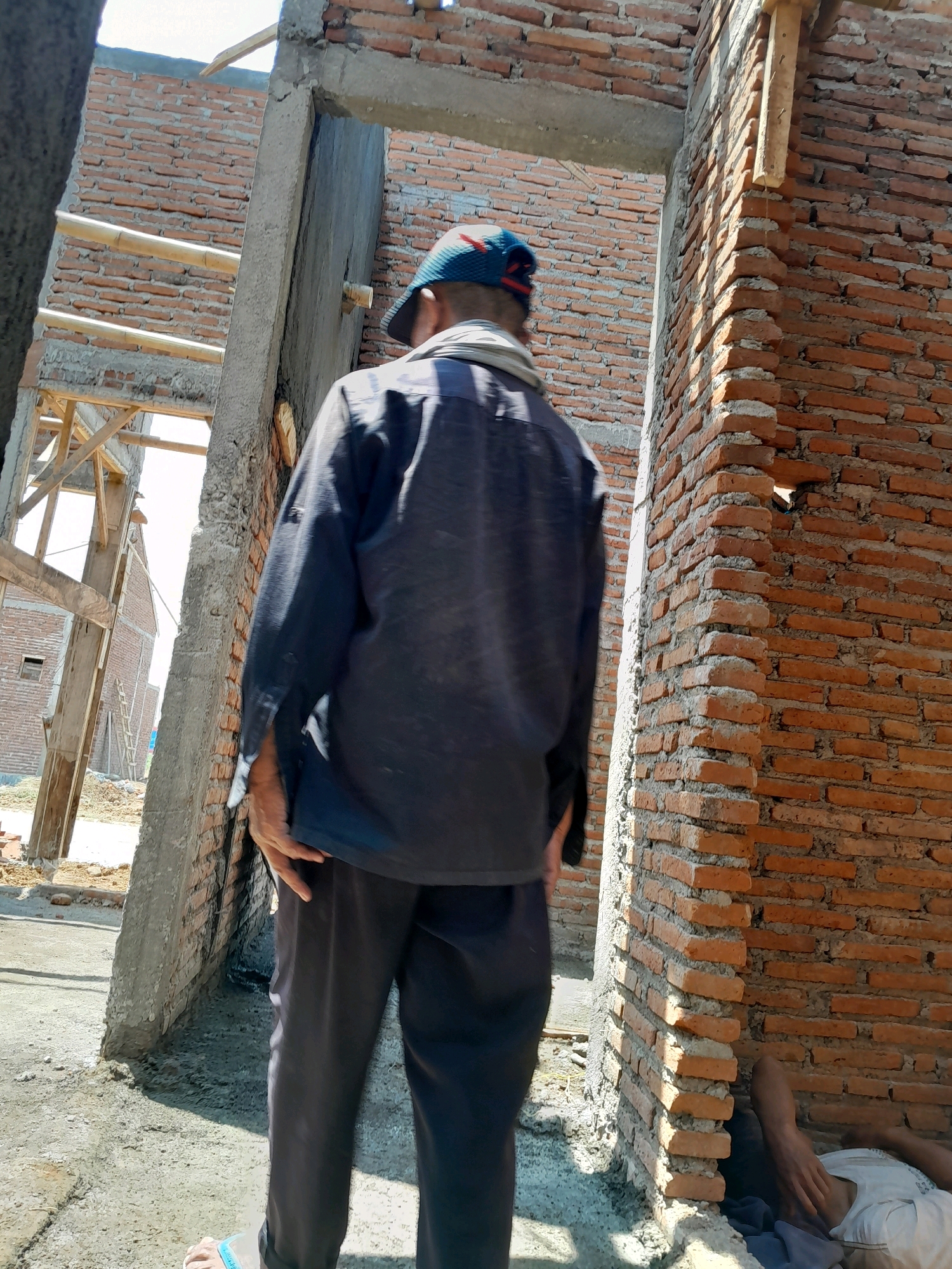 In Karangduren, Indonesia, at a construction site, a man wearing a dark shirt, dark pants, and a blue and red patterned hat stands facing away from the camera, looking into an unfinished doorway. To the right of the doorway, a brick wall is partially constructed. At the base of this wall, another individual is lying down, partially visible and wearing a white shirt. The ground is covered with concrete and rubble, suggesting ongoing construction. The background shows more unfinished brick structures and wooden scaffolding.