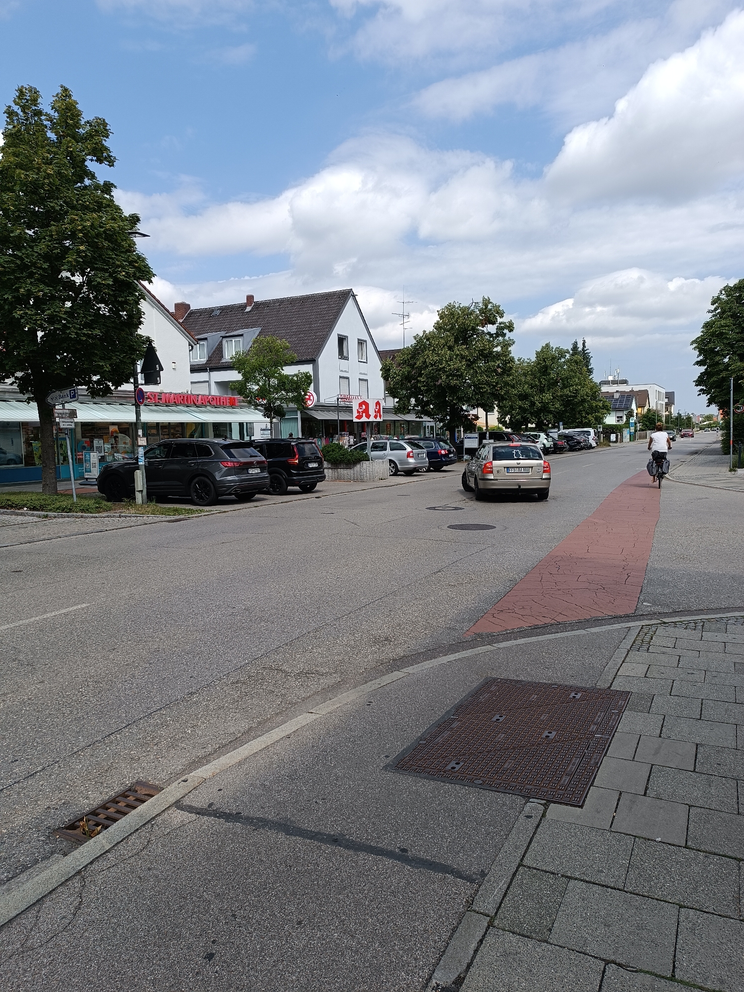 A street scene in Germering, Germany, features a road with parked cars and a cyclist. On the left side of the road are several businesses, including a pharmacy with a red "St. Martina Apotheke" sign and a "V. Bank P" sign. Several dark-colored SUVs are parked in front of the buildings. A silver sedan is in the foreground of the street, and a person on a bicycle is riding away from the camera on the right side of the road, on a red-marked lane. Trees line the sidewalk on the left, and buildings with pitched roofs are visible behind them. The sky is partly cloudy. A storm drain is visible in the lower left portion of the image, and a large metal manhole cover is on the sidewalk in the lower right.