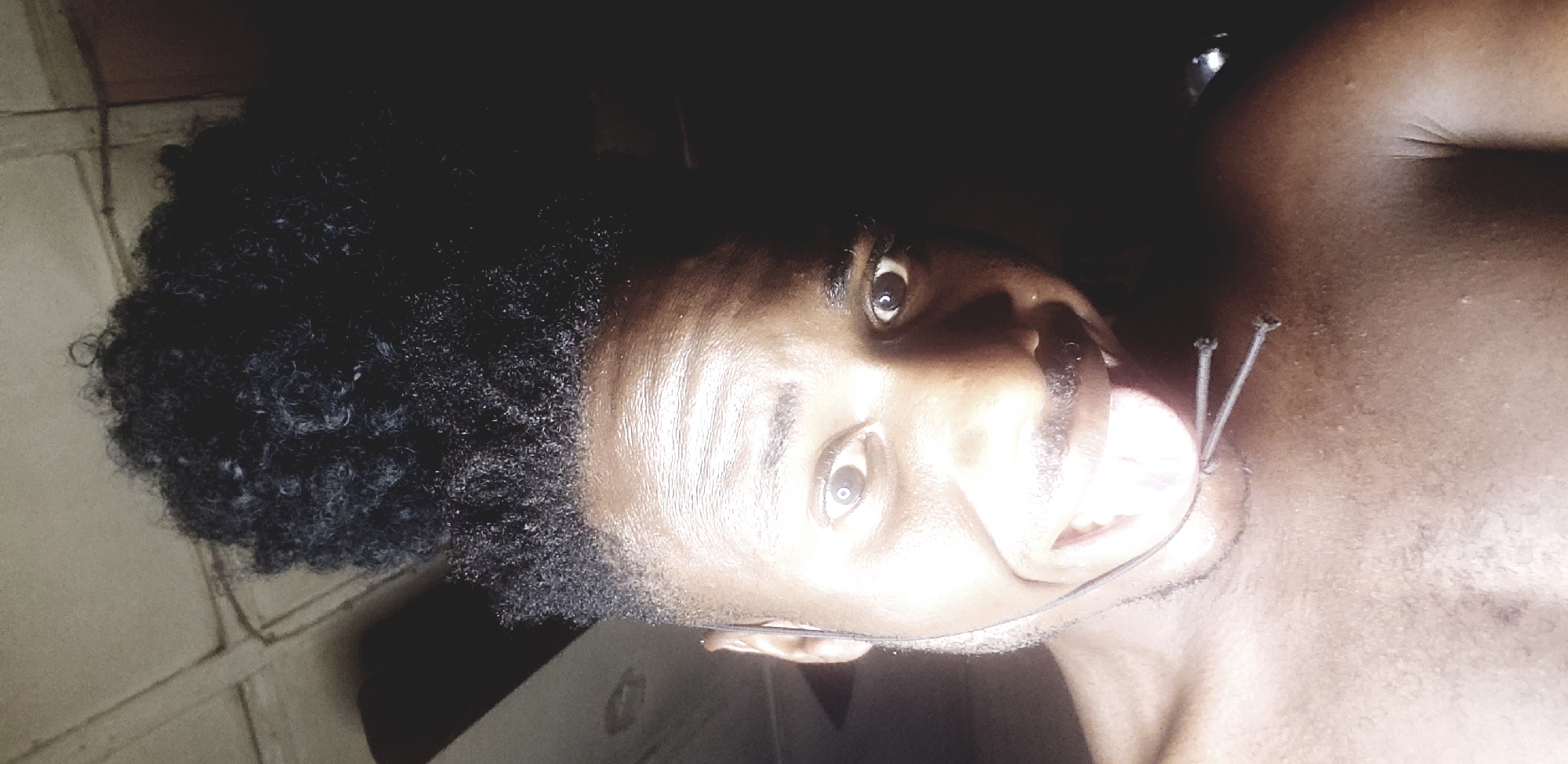 A person with dark skin and tightly curled, dark hair is lying on their back, facing upwards toward the camera. The person appears to have a dark stubble beard, wide eyes, and slightly parted lips. There are two pins stuck into the right side of their neck, above the collarbone. A thin black wire runs from near their ear, across their neck. The background includes a tiled floor on the left side and a dark, undefined space above and on the right. The image was taken in Narayi, Nigeria.