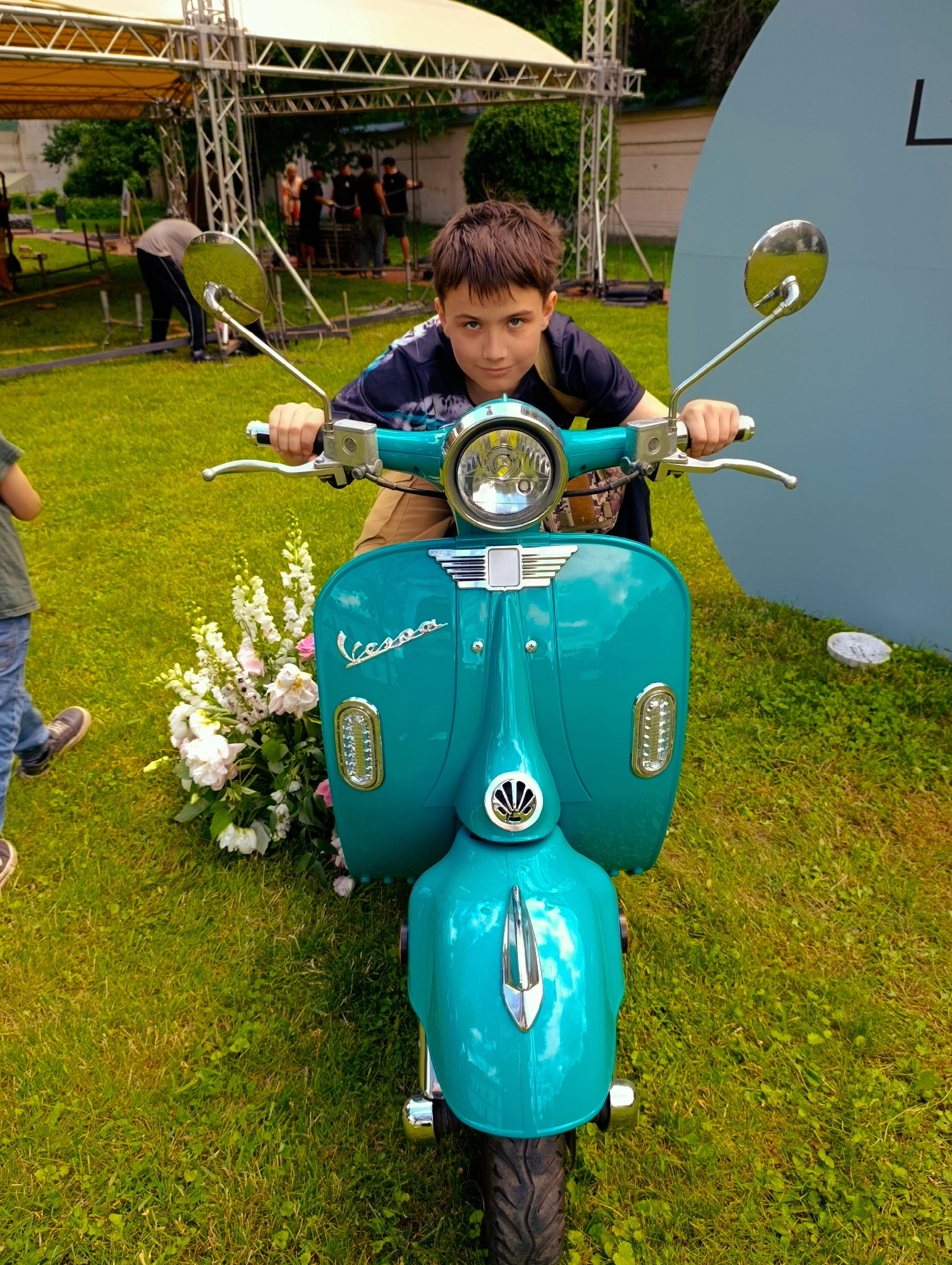 A young boy with short brown hair and blue eyes sits on a teal scooter. He is wearing a dark blue t-shirt with a tie-dye pattern on the sleeves and khaki shorts. The scooter is a classic Vespa model, with chrome accents and a prominent headlight. In front of the scooter, a bouquet of white and pink flowers is arranged on the grass. The scene takes place outdoors on a grassy area, with a large, light blue, circular structure and scaffolding in the background. Several people are visible in the background, some appearing to be setting up equipment. The overall impression is of a staged event or outdoor gathering. The image is taken from a low angle, looking up at the boy and the scooter. The lighting suggests it is daytime. The location is not identifiable from the image itself, and therefore the city of Kyiv, Ukraine cannot be determined.