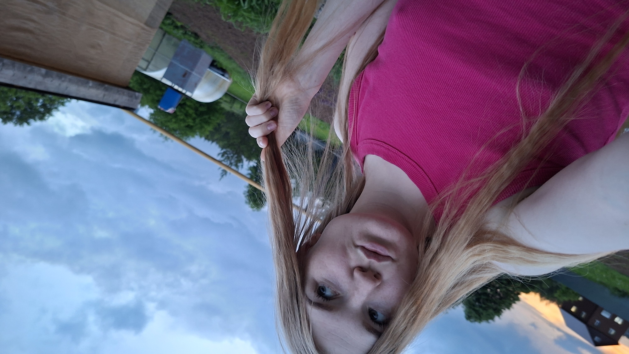 The media file is a selfie or self-portrait, presented rotated 90 degrees clockwise, featuring a person with long, light-colored hair, set against a rural or semi-rural backdrop. The person has fair skin and blue eyes, looking directly towards the camera with a neutral expression. They are wearing a vibrant fuchsia-colored, ribbed tank top or camisole. One hand is visible, gently holding a section of their hair near their chest.
The background elements are viewed as if the horizon runs diagonally from the lower-right to the upper-left of the rotated frame. A vast, cloudy sky dominates the lower-left portion, displaying shades of blue and grey, with hints of warmer light (orange/yellow) on some clouds towards the upper-right, suggesting either dawn or dusk. Above the horizon line, which appears to be the upper-right section of the image, there is cultivated land, likely a garden or field, with rich brown soil visible. Green foliage from trees is scattered across the mid-ground and distant background. A wooden pole or stick extends horizontally across the frame. In the mid-ground, a small, semi-circular greenhouse or tunnel structure is visible, alongside what appears to be a blue container. Further in the distance, a multi-story building with visible windows can be seen on the far right. The setting aligns with the provided location context of Большой Букор, Russia, suggesting a rural environment. No visible text is present in the image.